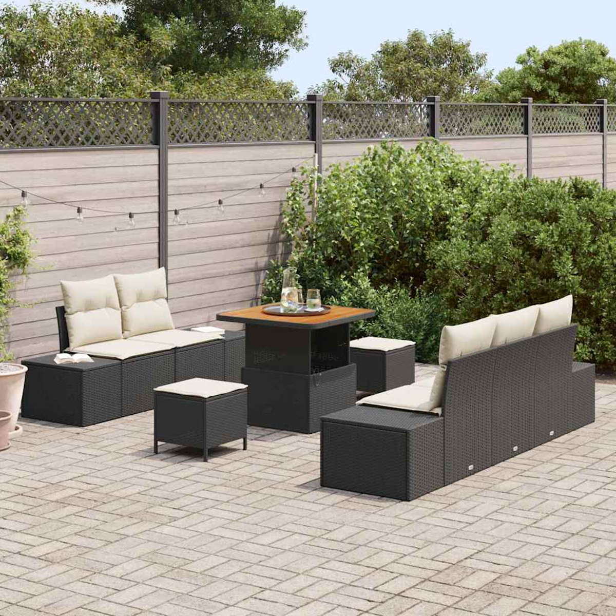 Ein Terrassenset mit Sofa, zwei Ottomanen und einem Tisch. Das Set ist dunkelgrau mit beigen Kissen. Der Hintergrund umfasst einen Zaun und Büsche.