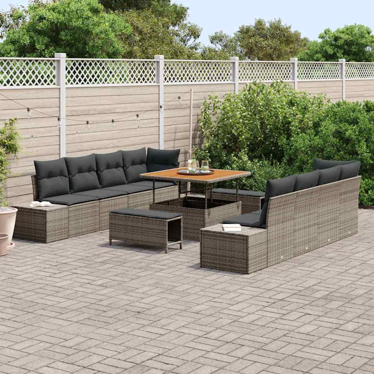 Gartenmöbel-Set. Graue Korbsessel, Tisch und Bänke mit dunkelgrauen Kissen auf einer Backstein-Terrasse, vor einem weißen Zaun.