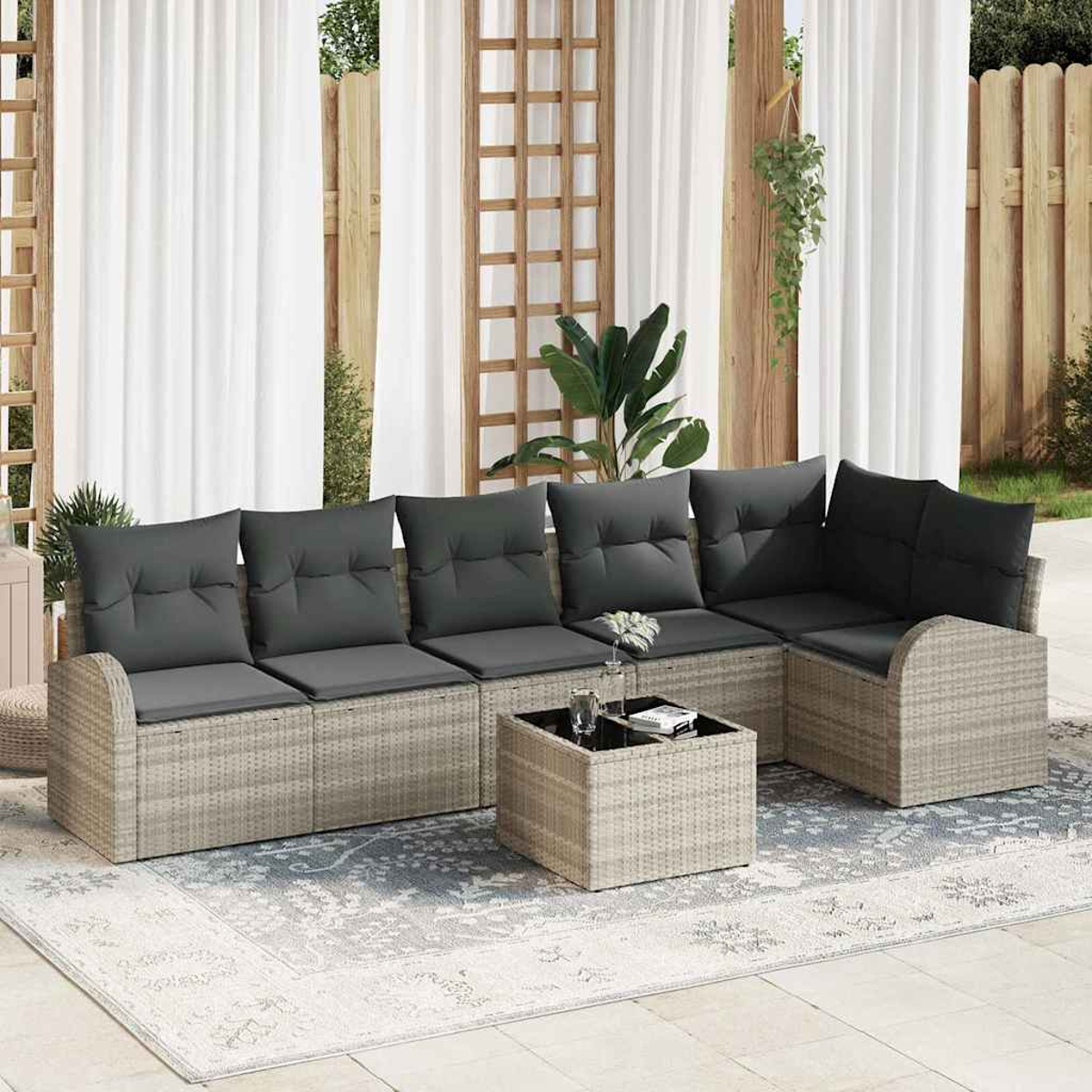 Outdoor-Ecksofa mit grauen Kissen auf einem gemusterten Teppich. Davor steht ein kleiner quadratischer Tisch. Weiße Vorhänge und ein Holzzaun im Hintergrund.