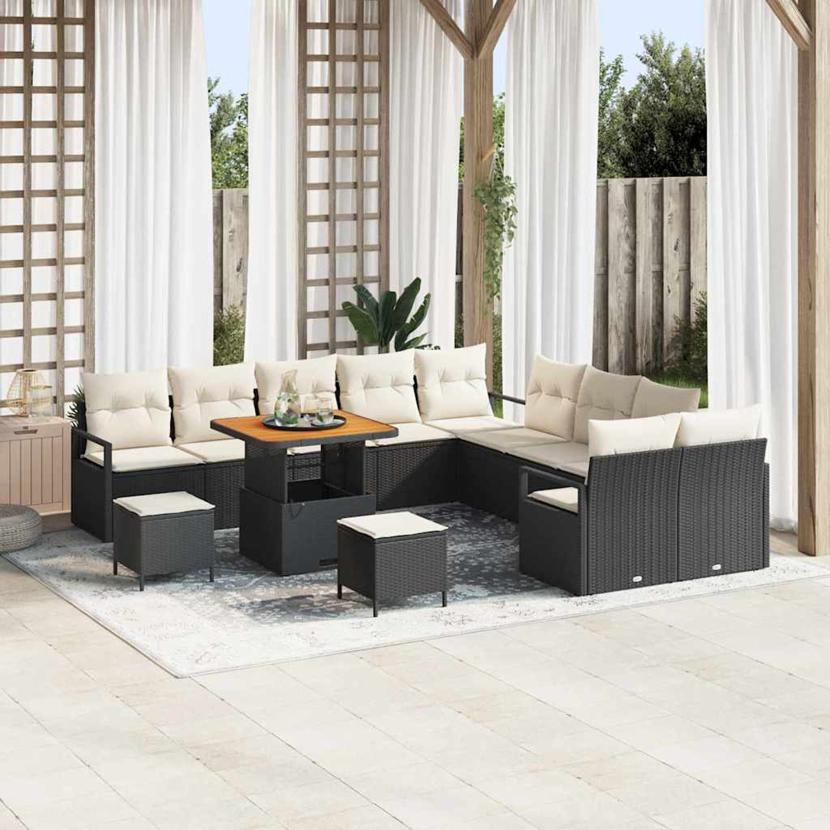 Gartenmöbel-Set mit schwarzen Korbsesseln, cremefarbenen Kissen, Holztisch und kleinen Hockern. Weiße Vorhänge und Grün im Hintergrund.