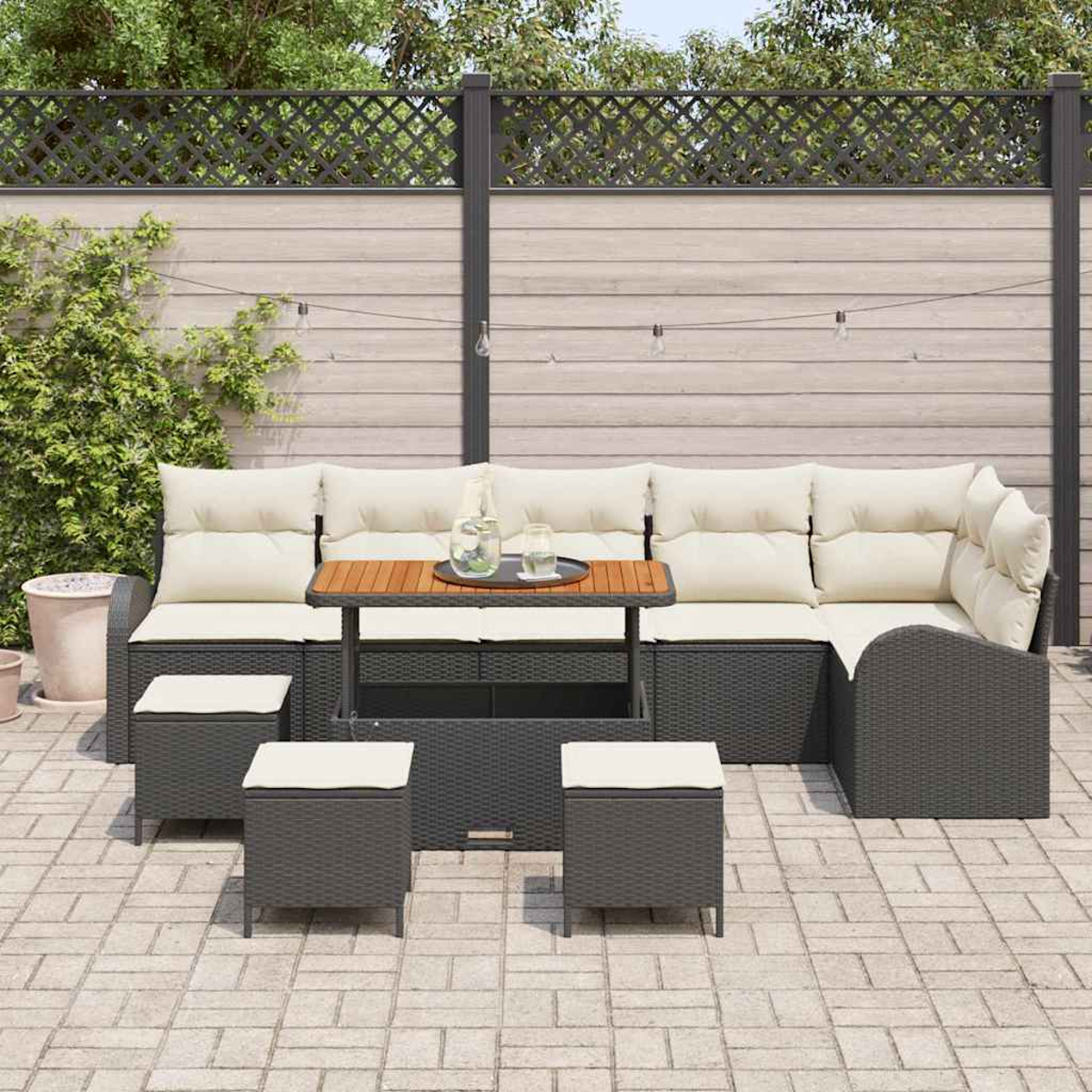 Set da patio esterno con divano, sgabelli e tavolo. Cuscini beige e vimini grigio scuro. Sfondo: recinzione, fogliame e patio in mattoni.