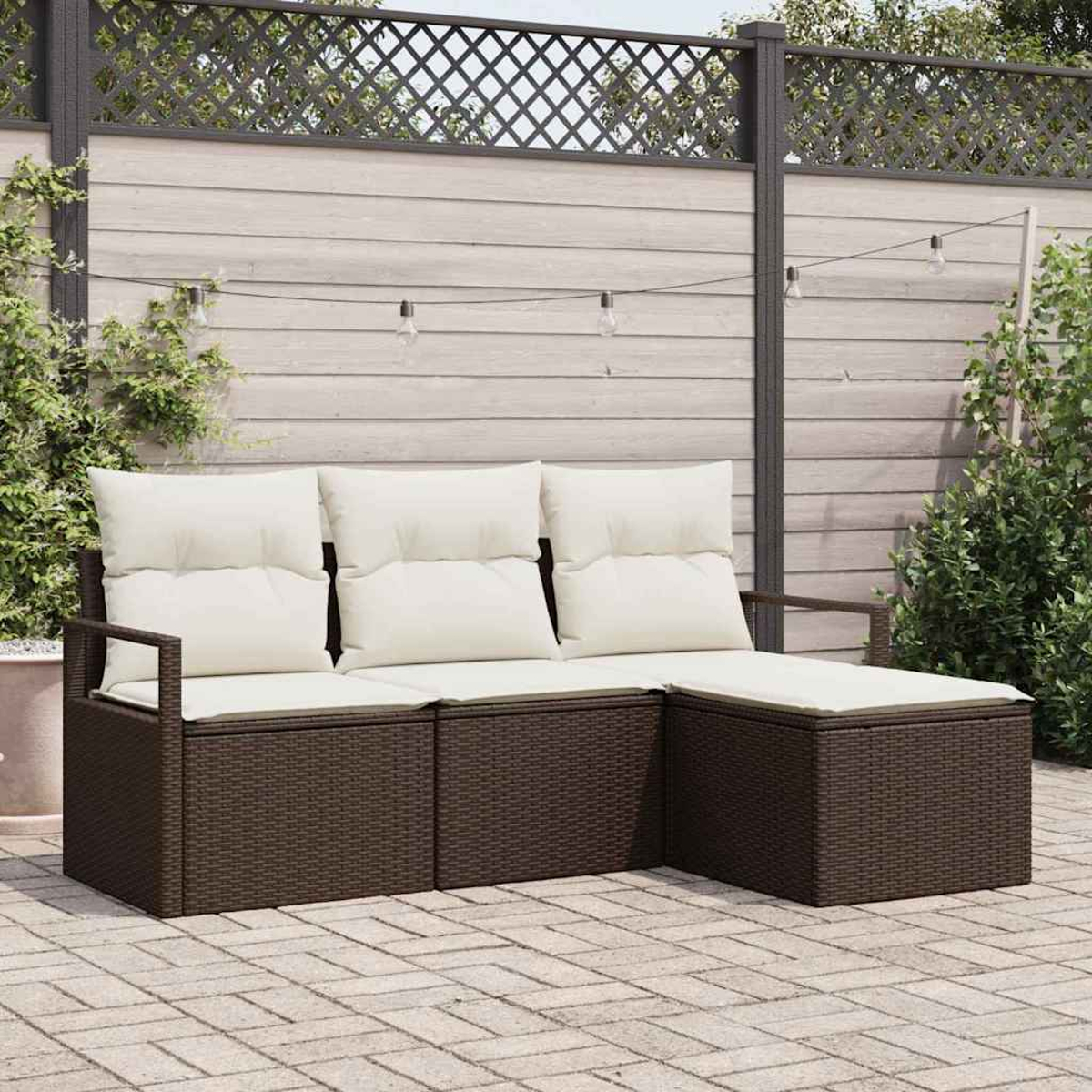 Ein braunes Outdoor-Sofa aus Korbgeflecht mit weißen Kissen. Im Hintergrund ein Holzzaun, Lichterketten und Grün.