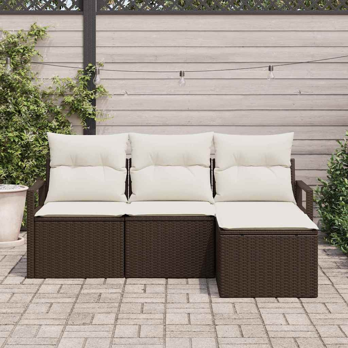 Ein braunes Rattan-Außensofa mit cremefarbenen Kissen steht auf einer Terrasse. Der Hintergrund zeigt einen Holzzaun und Grün.