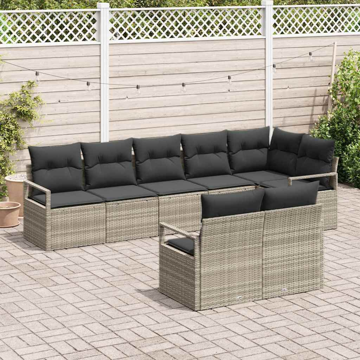Outdoor-Sitzarrangement. Beige Korbmöbel mit dunkelgrauen Kissen. Enthält ein Ecksofa und ein kleineres Sofa. Holzzaun-Hintergrund.
