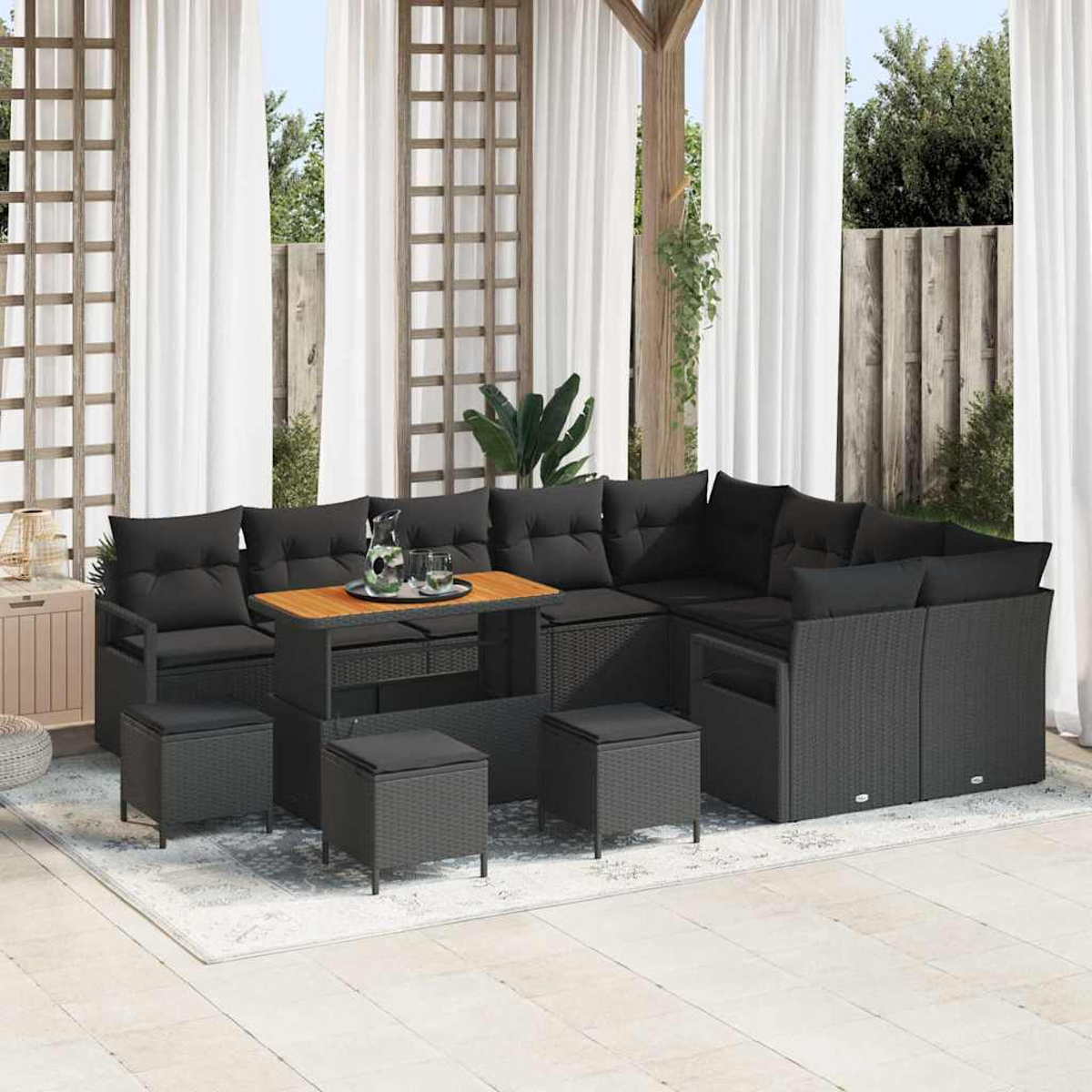 Garten-Terrassenset mit schwarzen Kissen, Holztisch und grauen Korbmöbeln. Weiße Vorhänge und ein Holzgitter im Hintergrund.