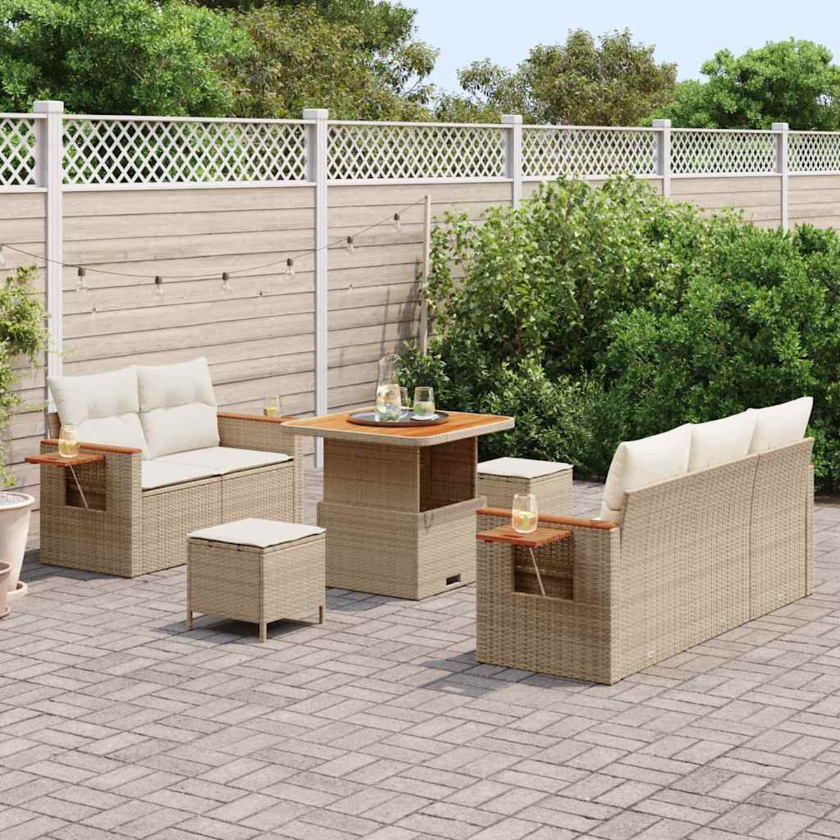 Set di sedute da esterno su un patio. Include divano, sedie, tavolo e pouf, tutti in vimini beige con dettagli in legno. Fogliame verde sullo sfondo.