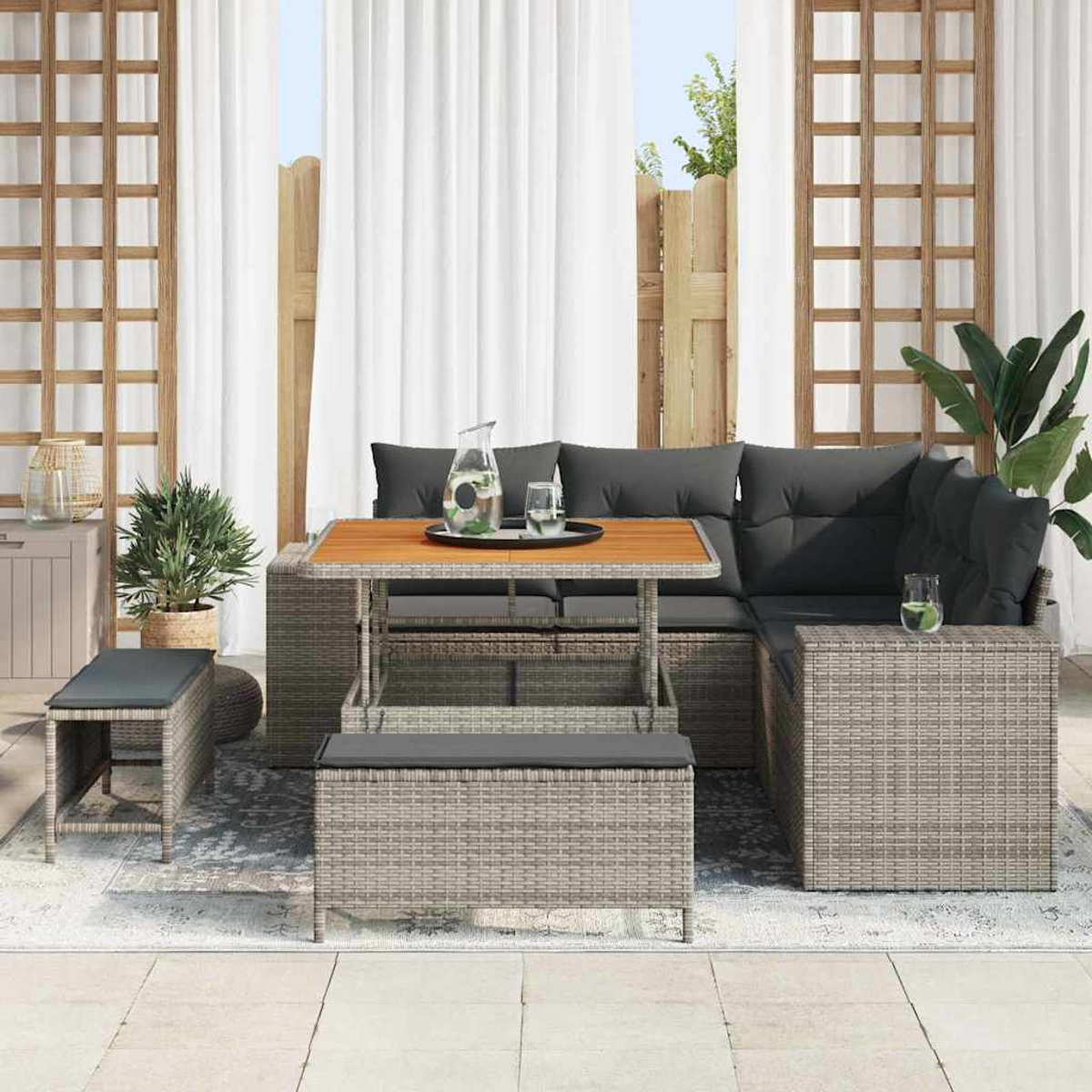 Gartensofa mit grauen Kissen, Holztisch und Bank. Weiße Vorhänge und Pflanzen im Hintergrund. Eine Kanne und Gläser stehen auf dem Tisch.