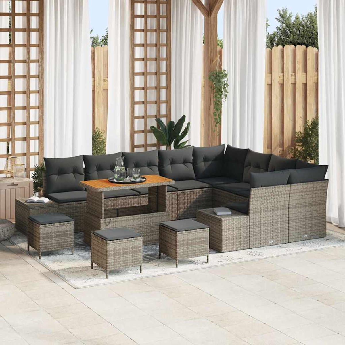 Gartenmöbel-Set. Graues Korbmöbel-Sofa, Couchtisch und Ottomanen auf einem Teppich, mit weißen Vorhängen im Hintergrund.