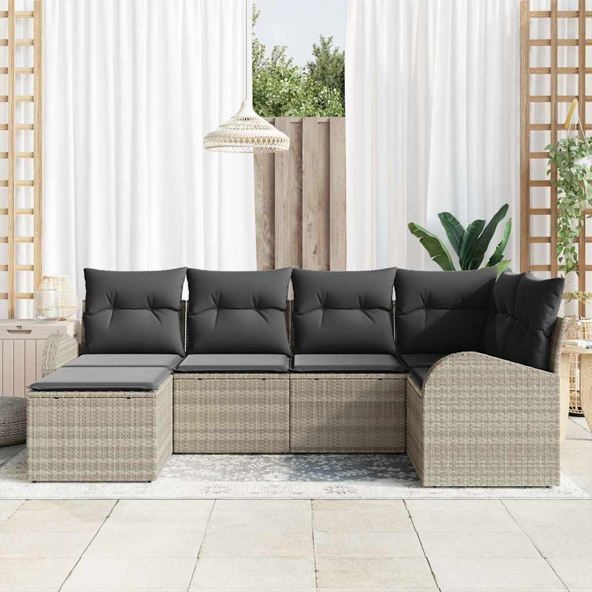 Geflochtenes Outdoor-Ecksofa mit grauen Kissen. Weiße Vorhänge, ein Fenster und Pflanzen im Hintergrund.