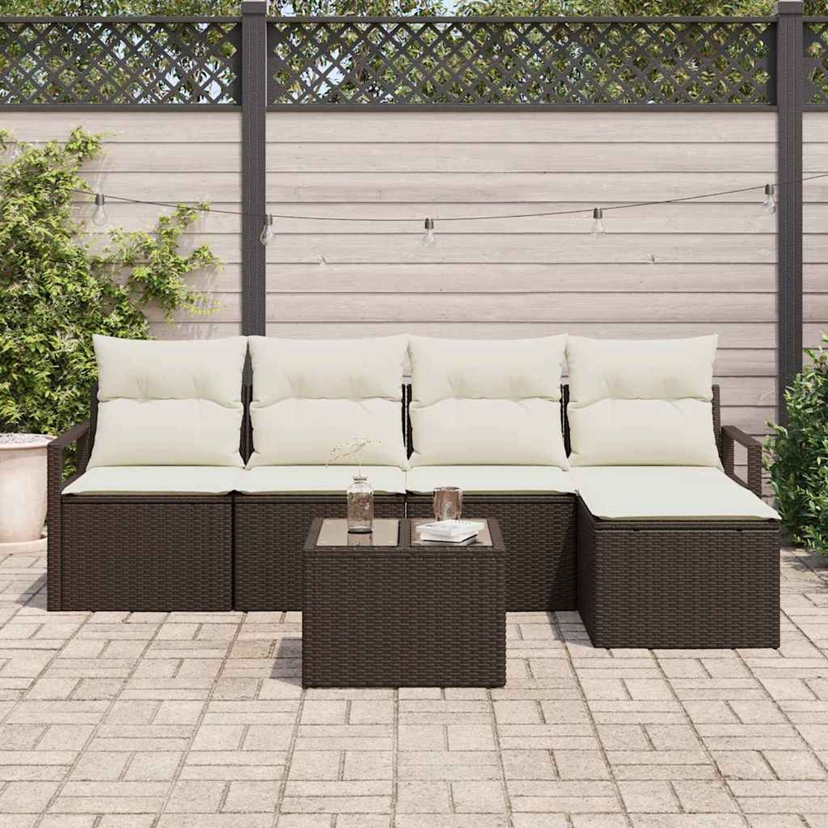 Gartenmöbel-Set. Dunkelbraunes, gewebtes Sofa mit cremefarbenen Kissen und einem kleinen Tisch auf einer gepflasterten Terrasse. Holzzaun im Hintergrund.