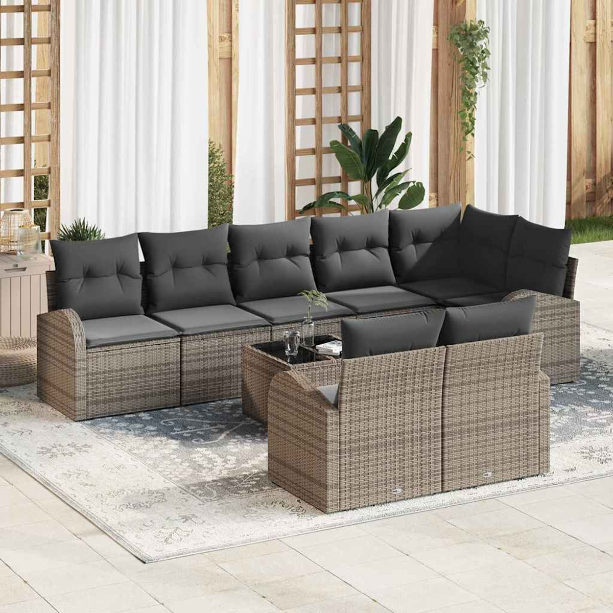 Ein graues Korbsessel-Ecksofa mit grauen Kissen steht auf einem Teppich. Ein Glastisch und Pflanzen befinden sich vor einem Vorhanghintergrund.