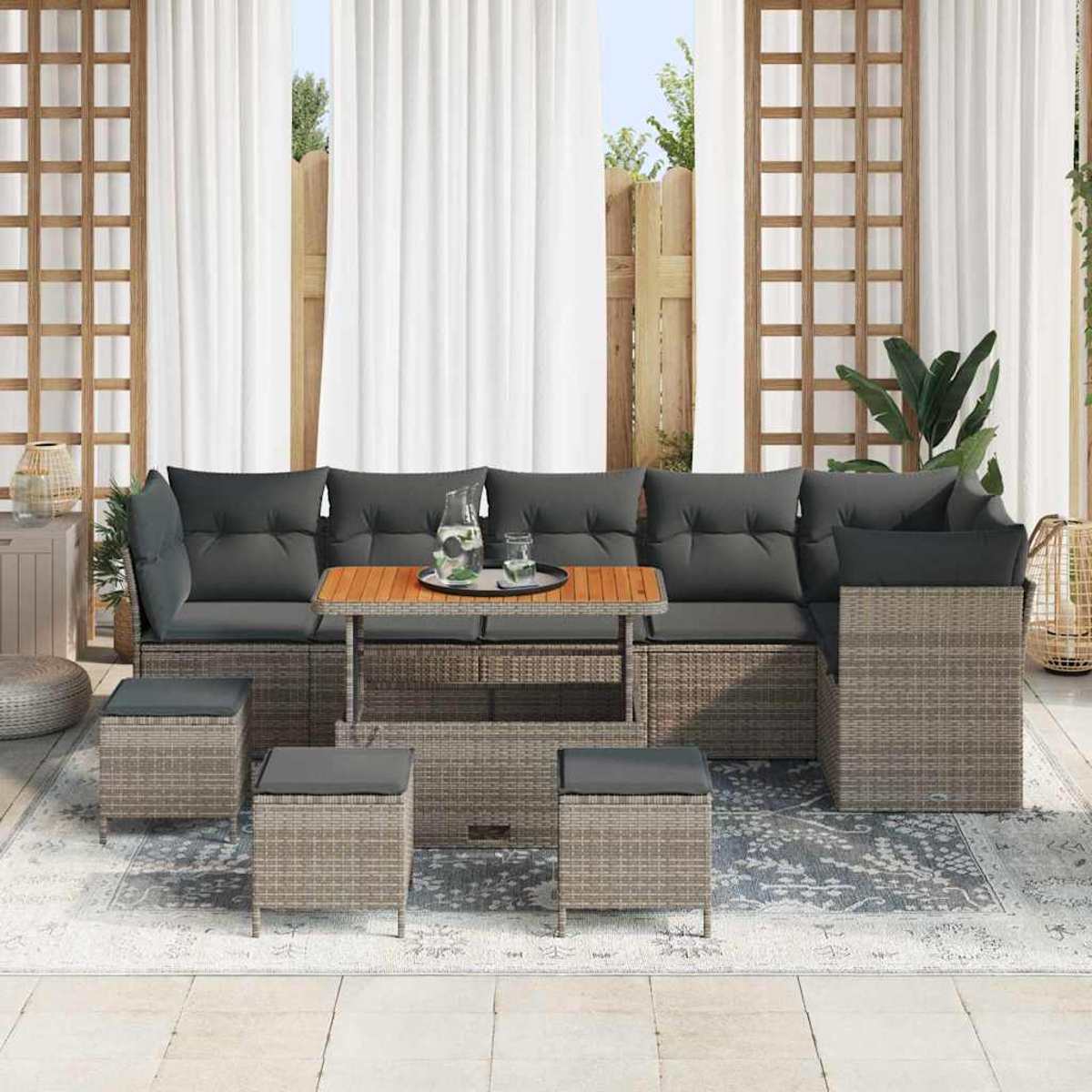 Gartenmöbel-Set: Sofa, Tisch und Hocker auf einem gemusterten Teppich. Weiße Vorhänge, Holzgitter und Pflanzen im Hintergrund.