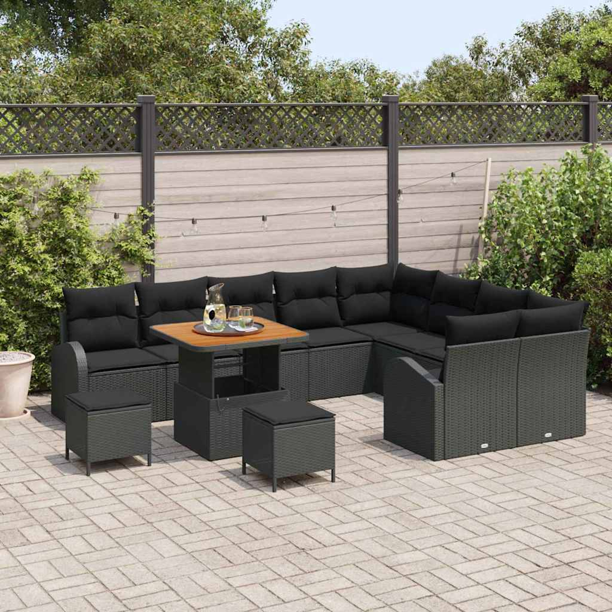 Terrassenmöbel: Schwarzes Korbsessel-Ecksofa mit Kissen, passenden Hockern und einem Tisch auf einer Ziegelterrasse vor einem Zaun.