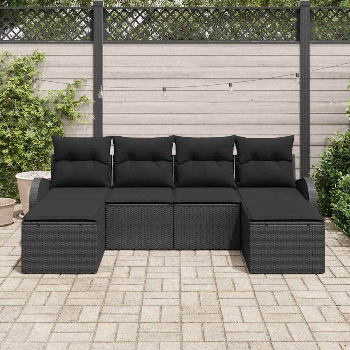 Ein schwarzes Outdoor-Ecksofa steht auf einer Steinterrasse. Dahinter befindet sich ein Holzzaun mit dekorativer Spitze und Lichterketten.