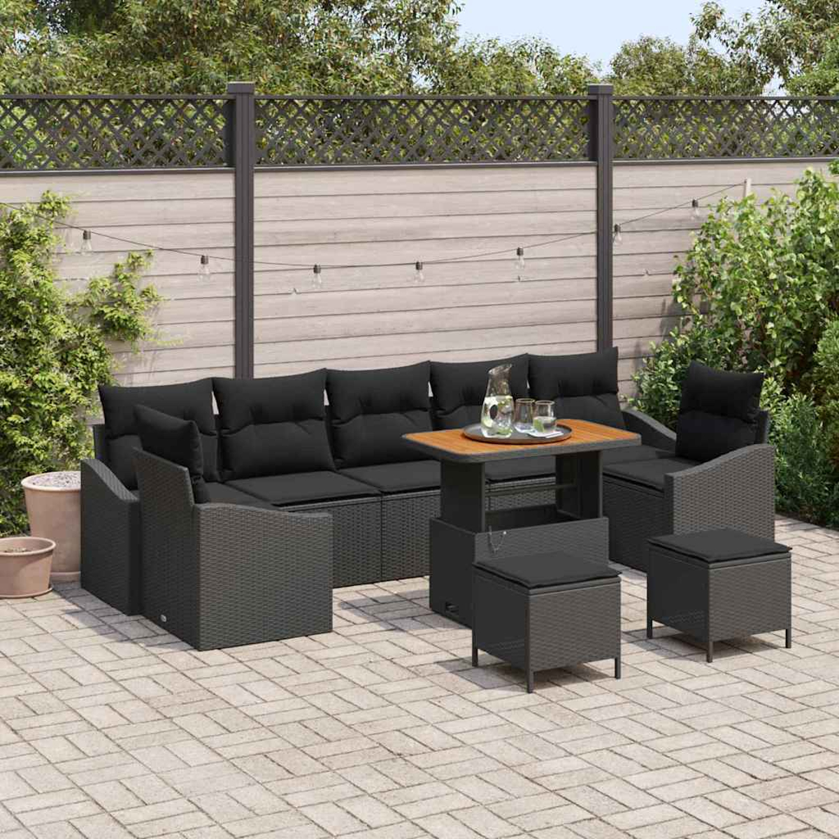 Terrassen-Set mit schwarzen Kissen, einem Couchtisch und zwei Ottomanen. Das Set steht auf einer Backsteinterrasse mit Pflanzen im Hintergrund.