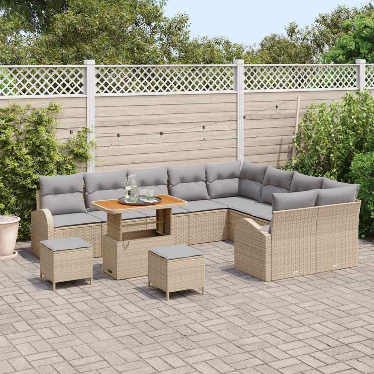 Ein Ecksofa im Freien mit grauen Kissen und einem Holztisch, zwei Ottomanen auf einer gepflasterten Terrasse. Ein weißer Zaun und grüne Pflanzen im Hintergrund.