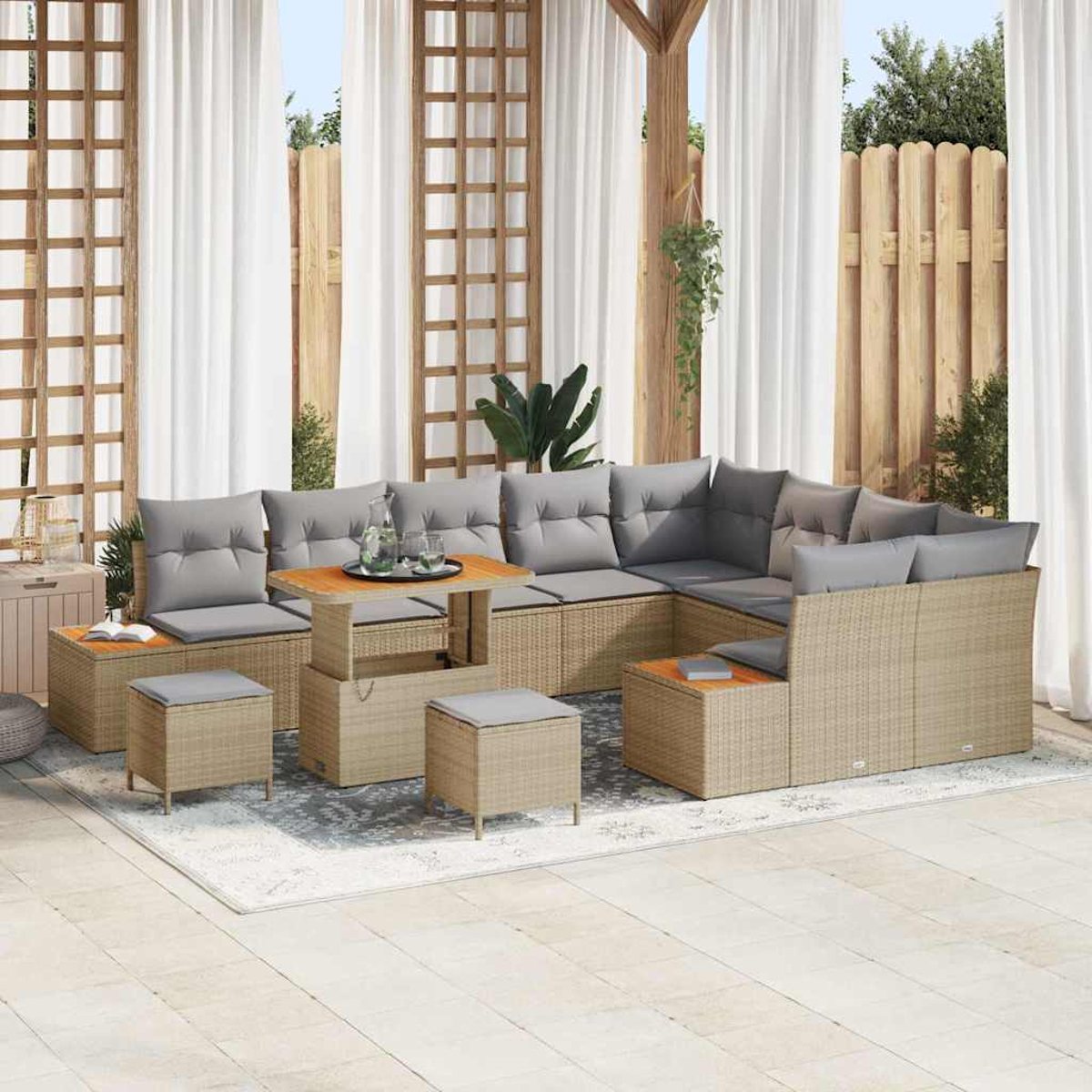 Zona de estar al aire libre con muebles de mimbre, cojines grises, mesa de madera y cortinas blancas translúcidas. Fondo con valla de madera y vegetación.