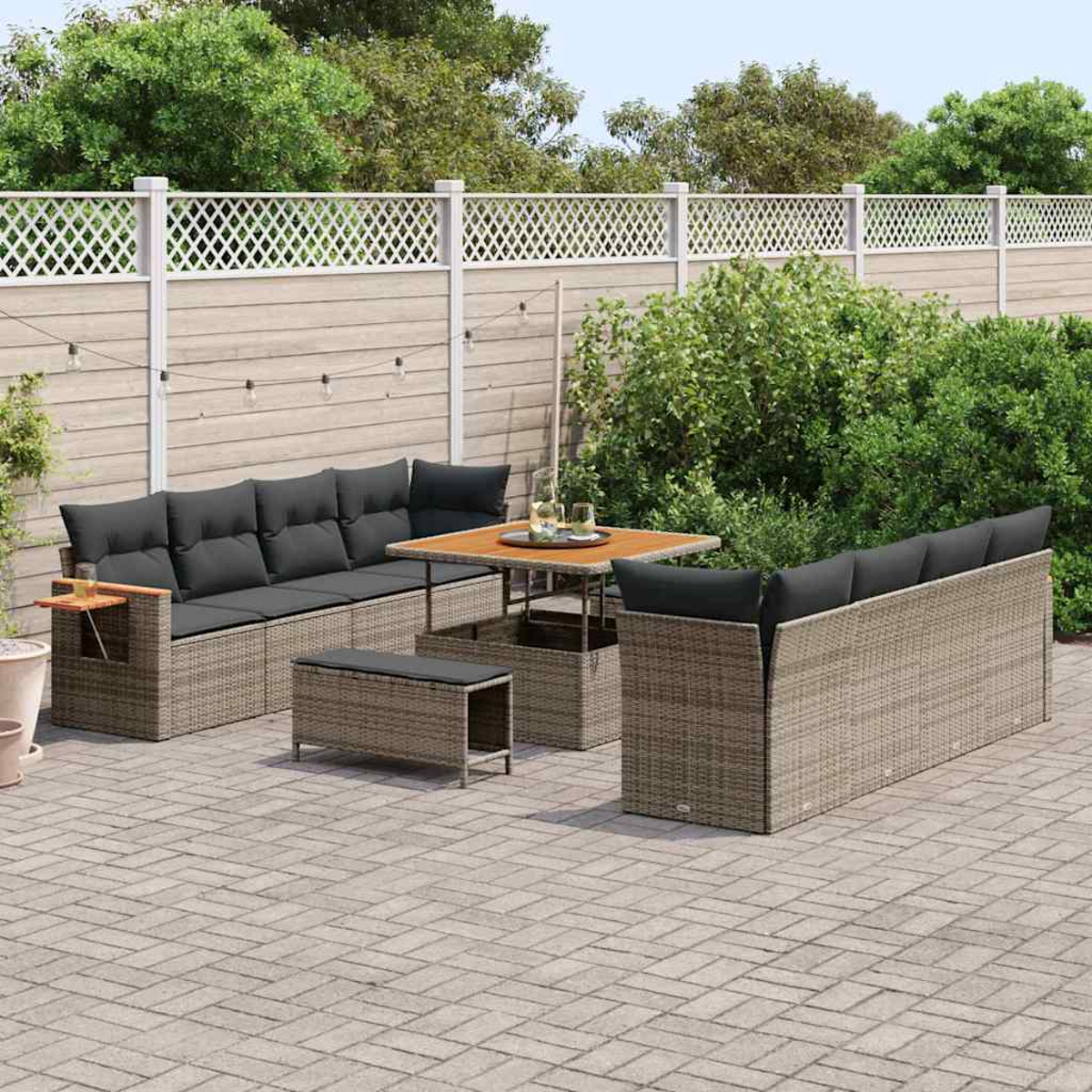 Set da patio esterno con cuscini grigi, tavolo in legno e mobili intrecciati, contro una recinzione a traliccio bianca e fogliame verde.