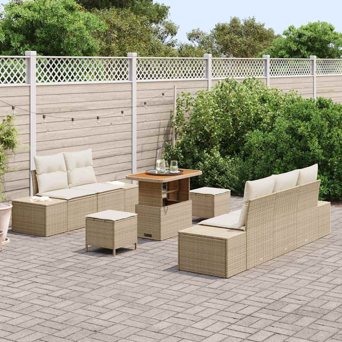 Ein Terrassenset mit beigen, gewebten Möbeln, darunter ein Sofa, eine Ottomane, ein Tisch und Hocker. Vor einem Zaun und Grün.
