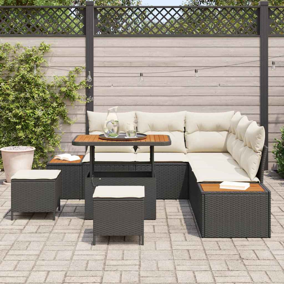 Gartenmöbel-Set mit L-förmiger Couch, Hockern und Tisch. Cremefarbene Kissen und eine Tischplatte aus Holz vor einem Zaun.