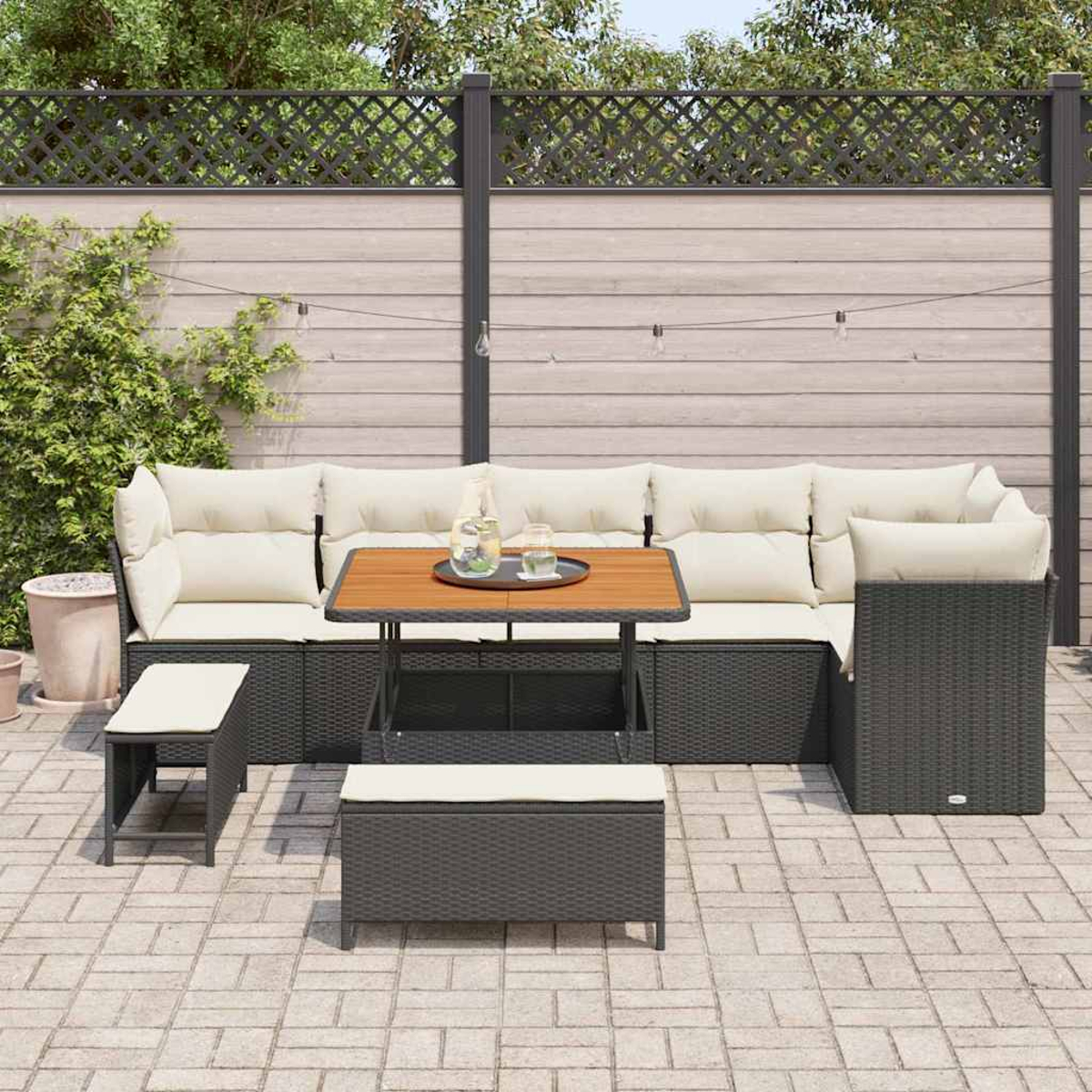 Eine Terrassengarnitur mit Sofa, Tisch und Bänken. Beige Kissen auf dunklen Korbmöbeln. Holzzaun im Hintergrund.