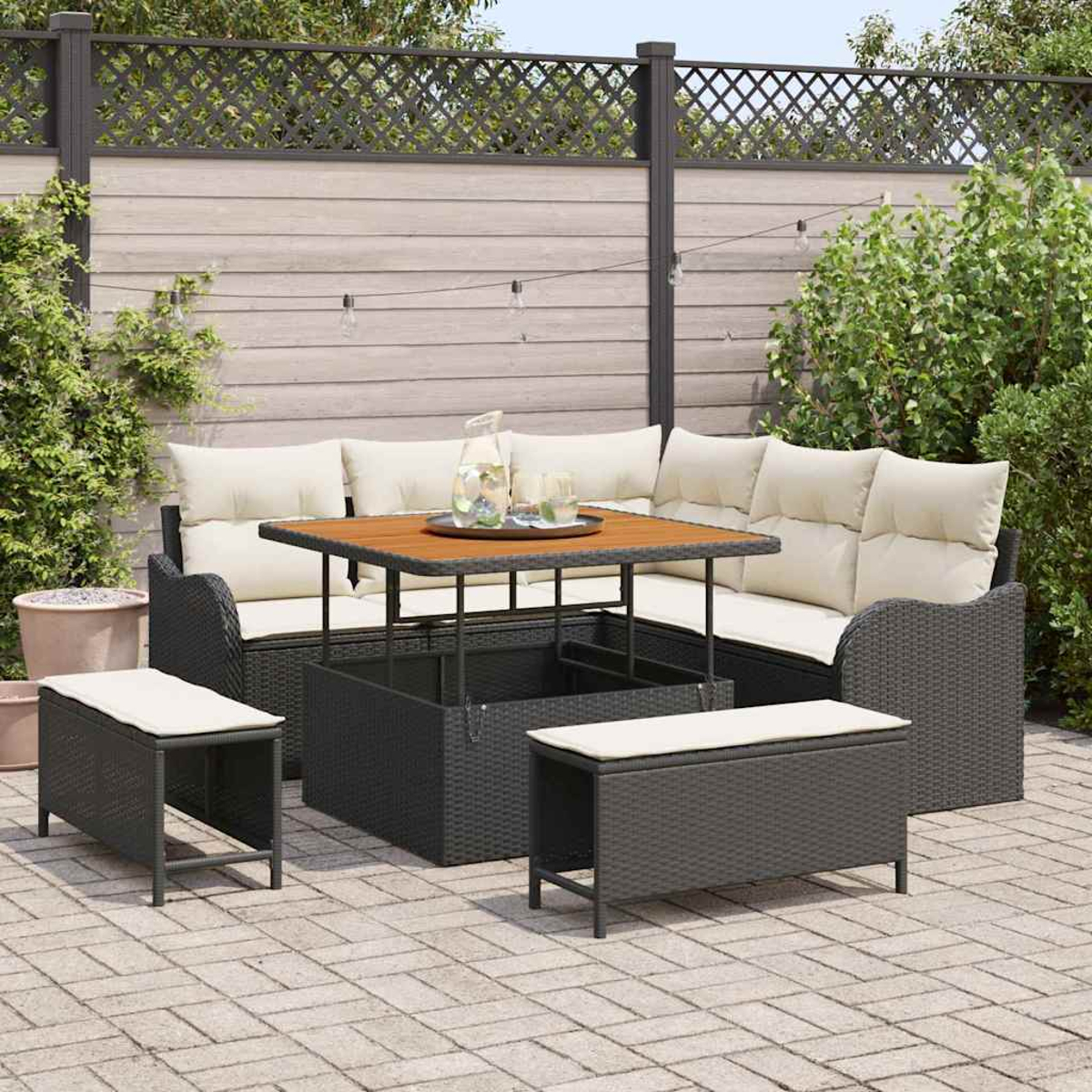Gartenmöbel-Set auf einer Backsteinterrasse. Es umfasst ein Ecksofa, einen Tisch und zwei Bänke, alles dunkles Korbgeflecht mit cremefarbenen Kissen.