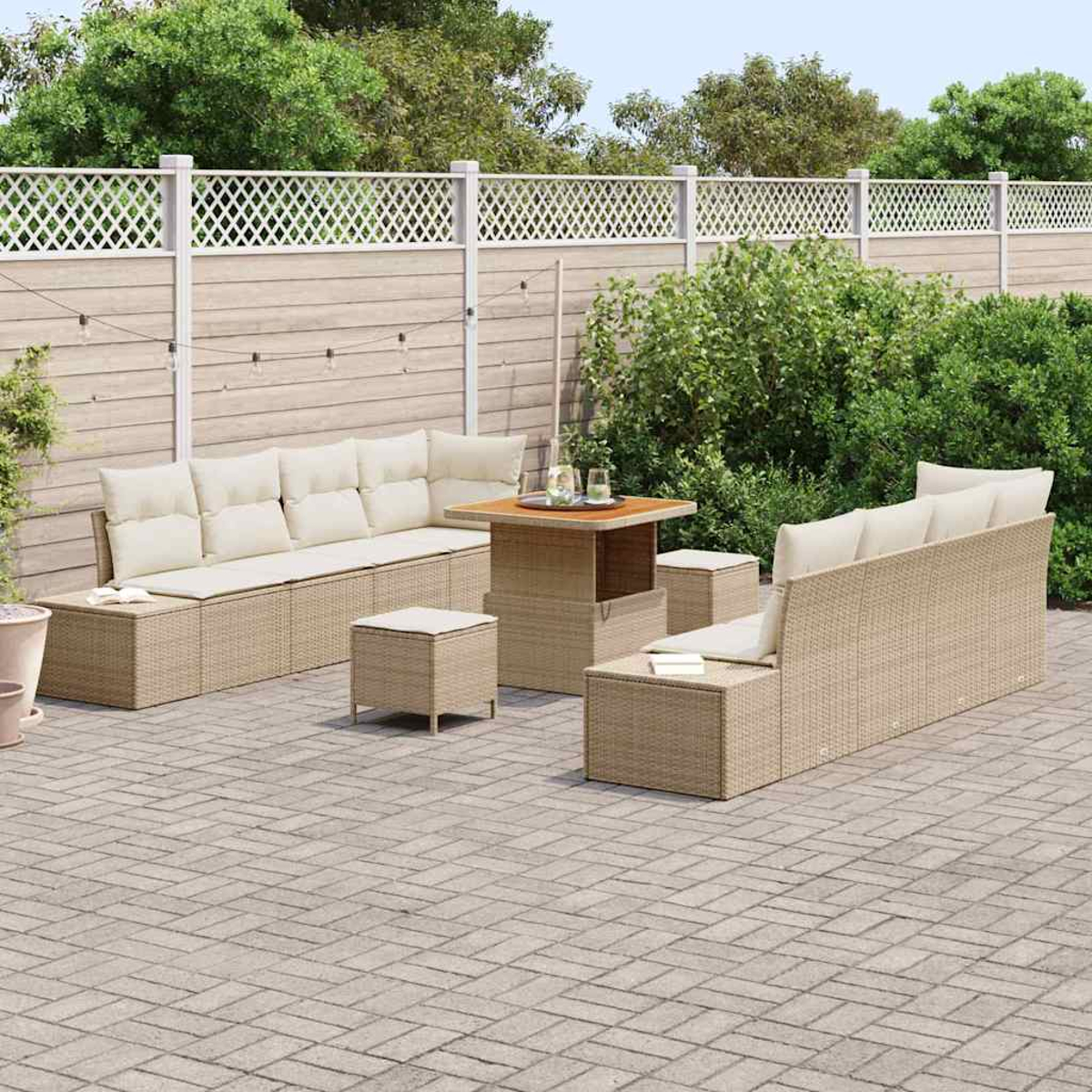 Garten-Sitzgruppe. Beige Korbsessel, Tisch und Hocker auf einer Backstein-Terrasse, dahinter ein Zaun und Grün.