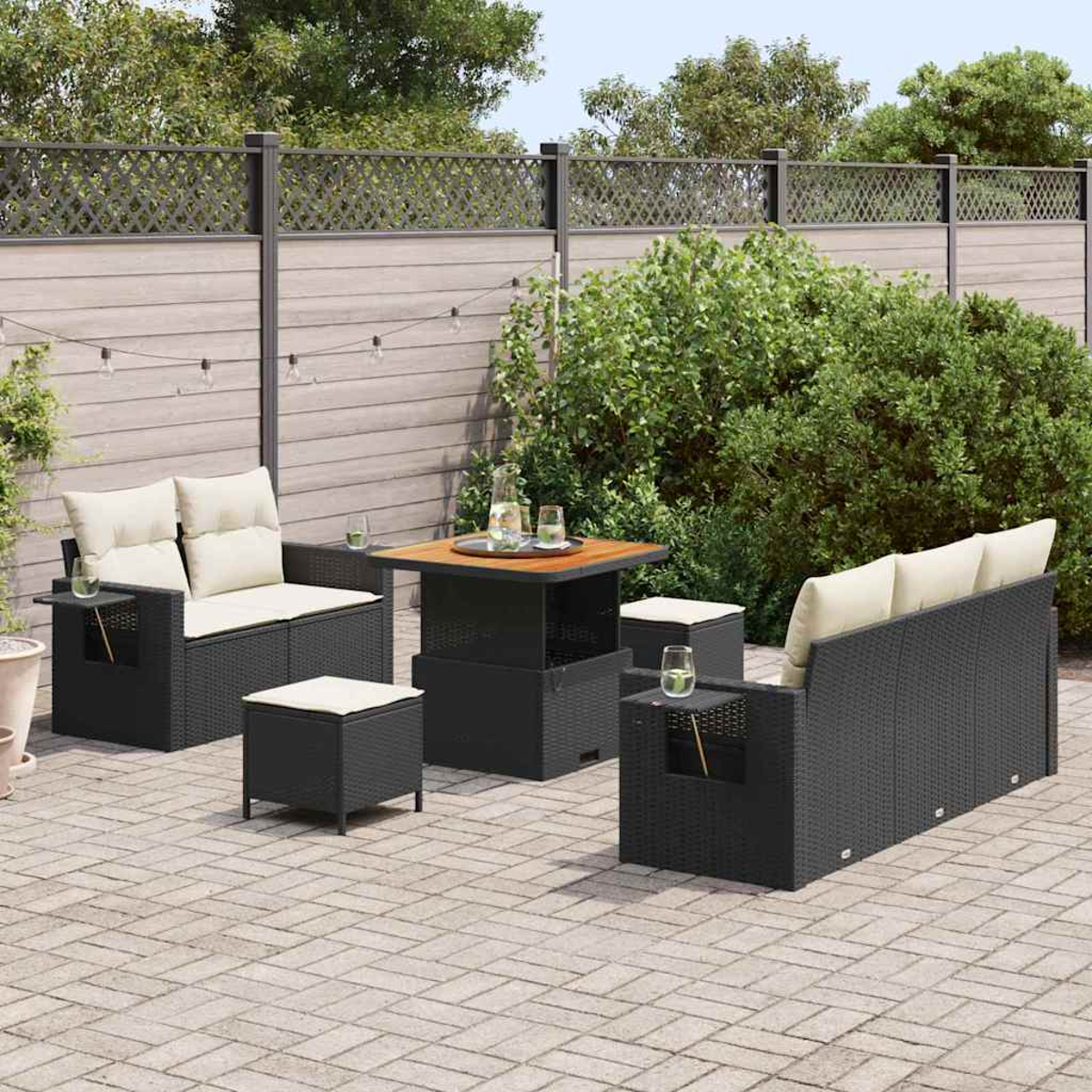 Terrassenset mit schwarzen Korbmöbeln, beigen Kissen und einem Holztisch. Im Hintergrund ein Zaun und Grün.