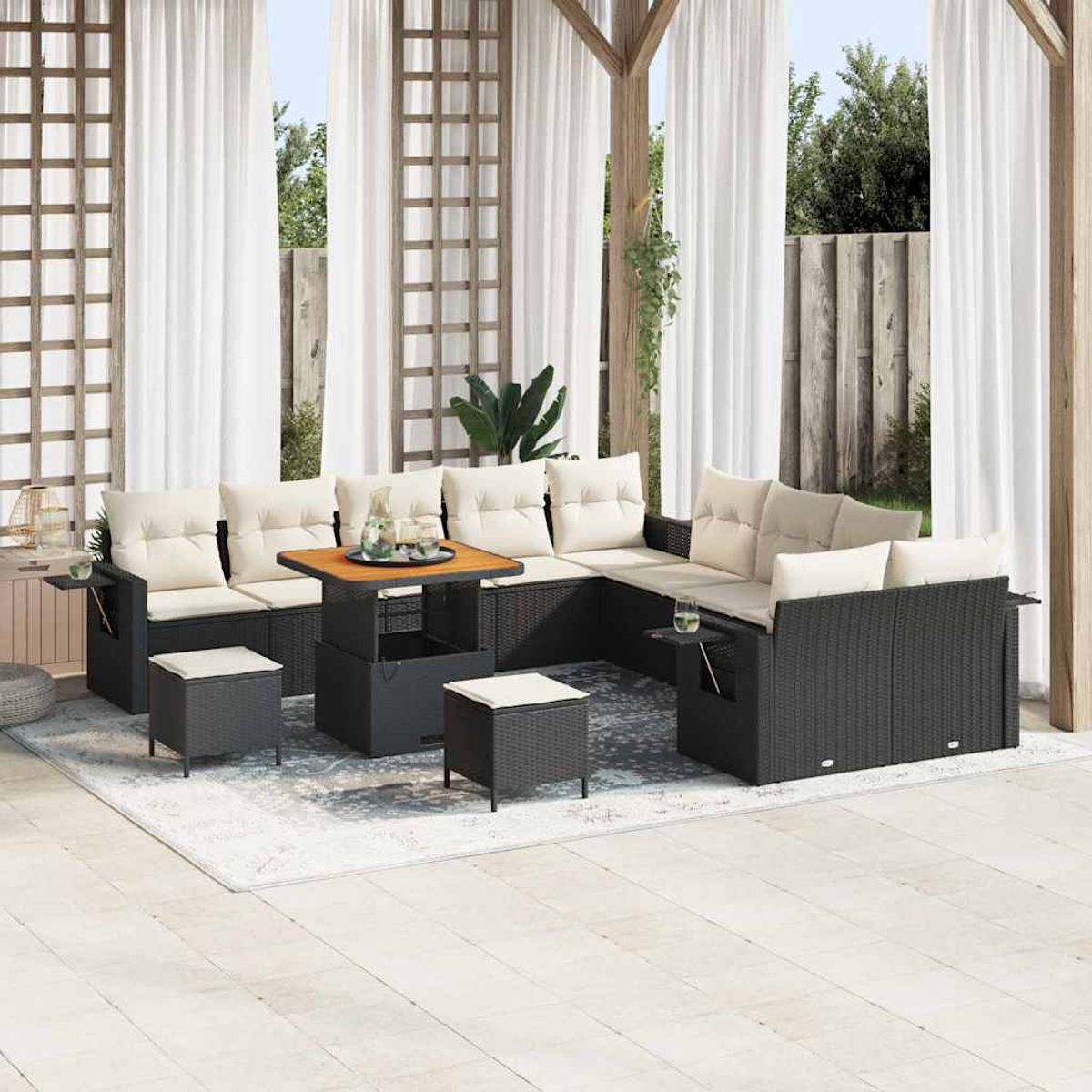 Juego de patio al aire libre con muebles de mimbre negro, cojines beige y una mesa de madera. Cortinas blancas y una alfombra definen el espacio.