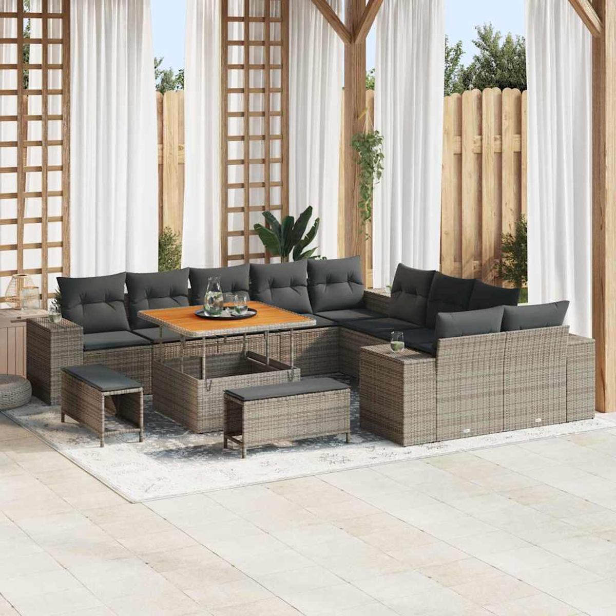 Außenbereich mit Rattansofa, Holztisch, grauen Kissen und weißen Vorhängen. Pflanzen und Holzzaun im Hintergrund.