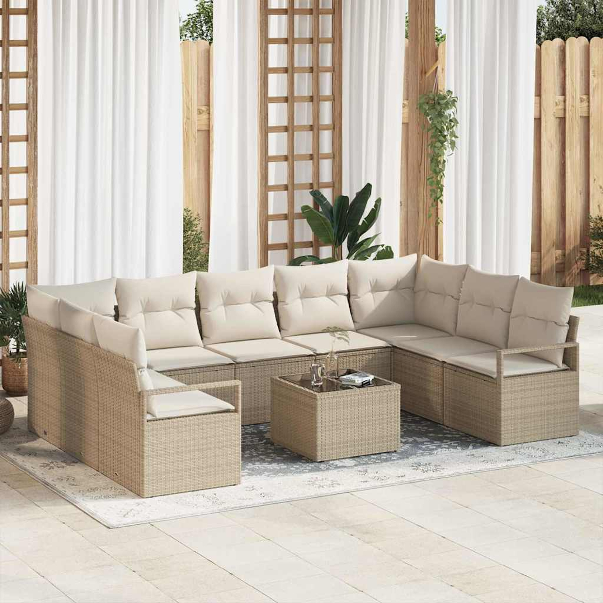 Outdoor-Korbsofa mit cremefarbenen Kissen und einem quadratischen Couchtisch auf einem gemusterten Teppich. Weiße Vorhänge und Holzzaun im Hintergrund.