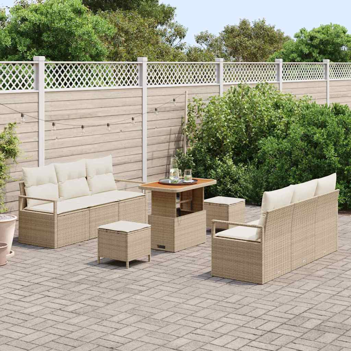 Gartenmöbel-Set aus Rattan. Beige Sofas und Ottomanen mit cremefarbenen Kissen. Holztisch mit Gläsern. Grüne Hecke und weißer Zaun im Hintergrund.