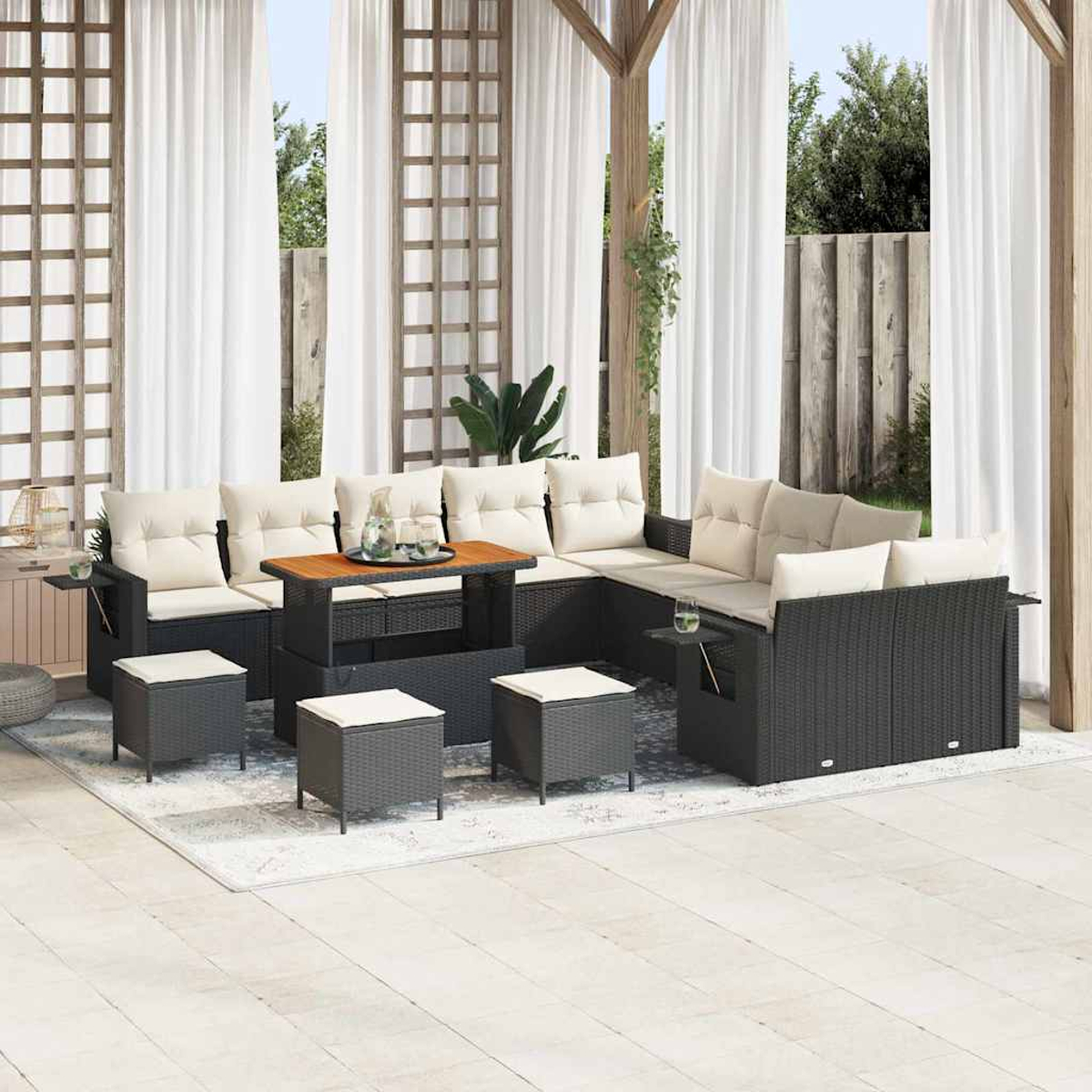 Outdoor-Sitzbereich mit schwarzen Rattanmöbeln, beigen Kissen, einem Holztisch und weißen Vorhängen. Hintergrund umfasst Pflanzen und Holzstruktur.