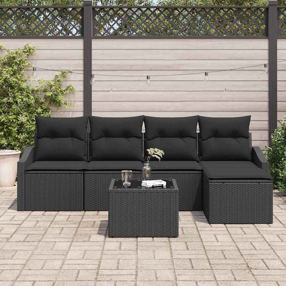 Schwarzes Outdoor-Wicker-Sofaset mit Kissen, kleinem Glastisch und Pflanzen. Holzzaun im Hintergrund, Lichterketten.