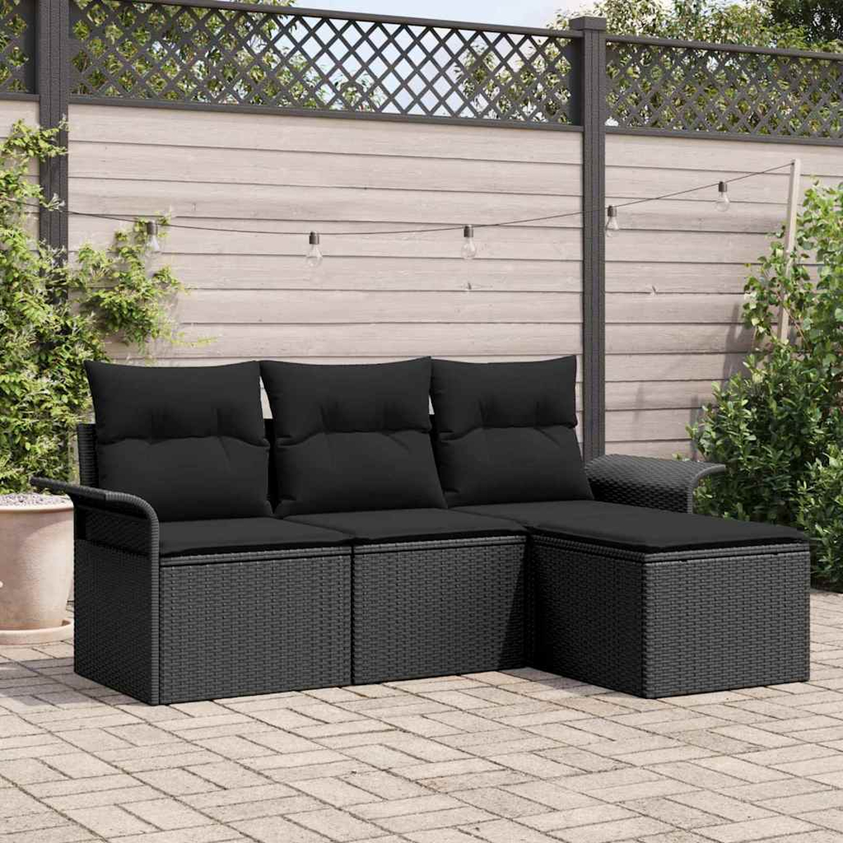 Schwarzes Outdoor-Ecksofa aus Korbgeflecht mit Kissen auf einer Terrasse. Im Hintergrund befinden sich ein Holzzaun und Lichterketten.