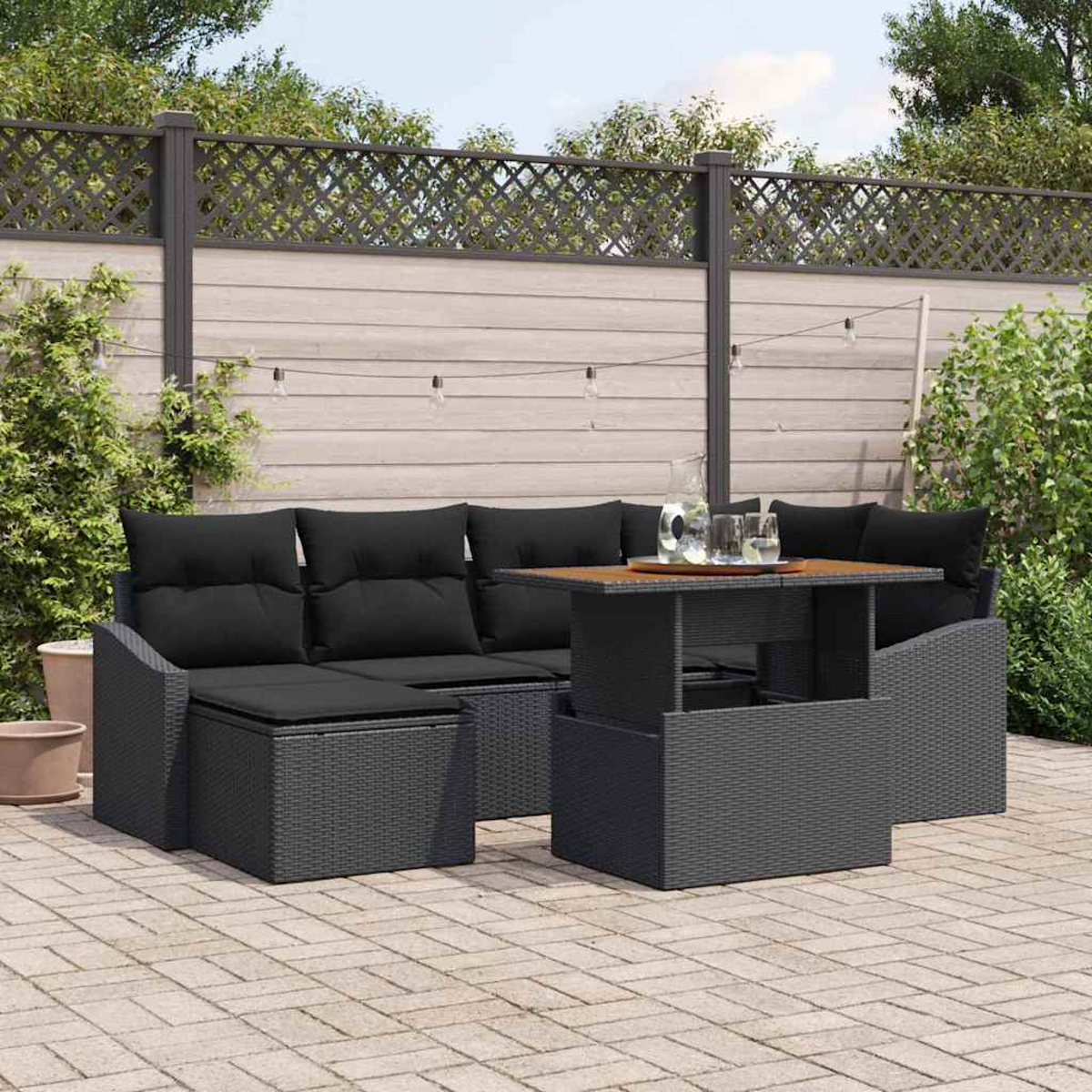 Terrassenset mit schwarzen Kissen und Tisch. Auf einer Backstein-Terrasse mit Zaun und Grün im Hintergrund.