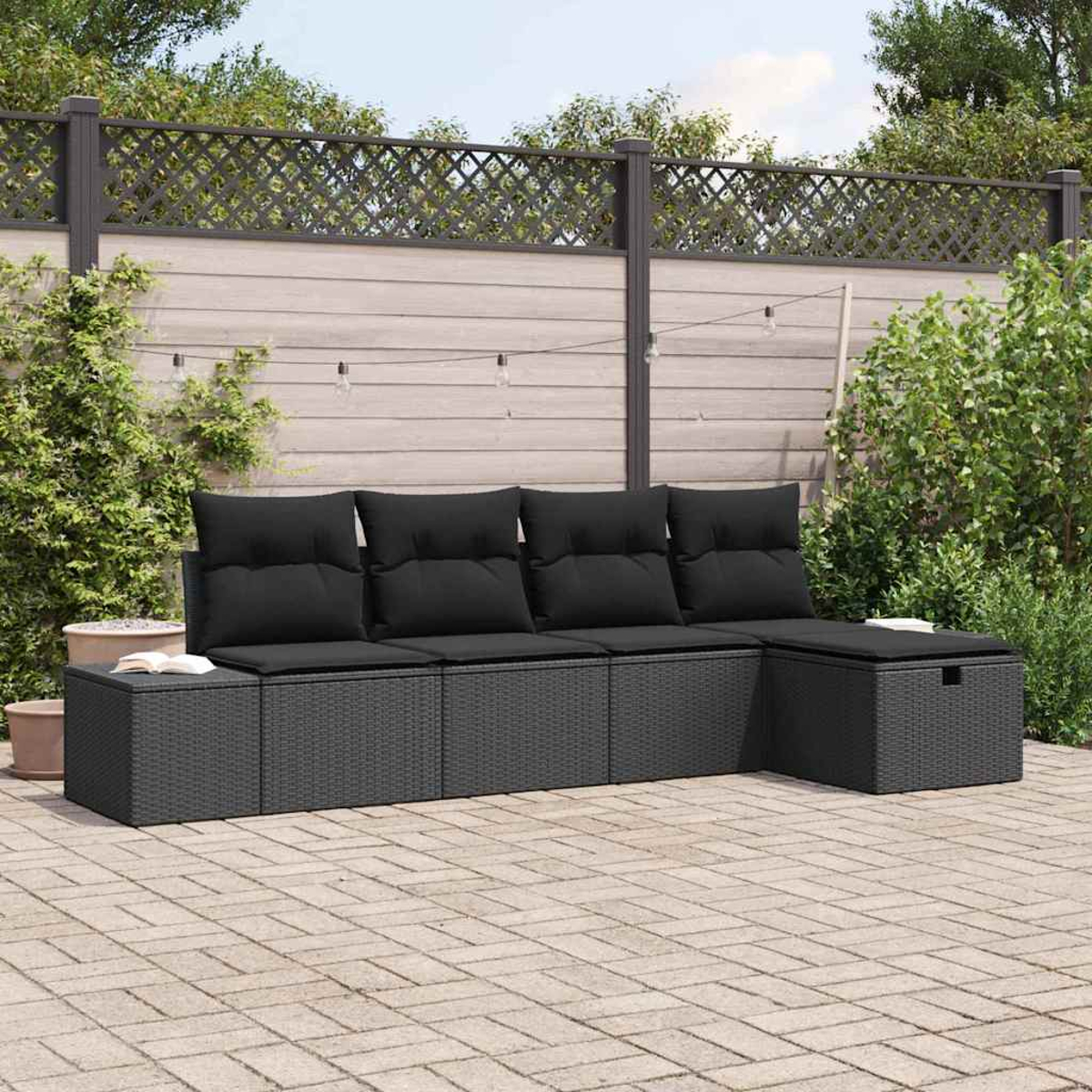 Ein graues Rattan-Sofa mit schwarzen Kissen steht auf einer Ziegelterrasse. Im Hintergrund ein Holzzaun und Grün.