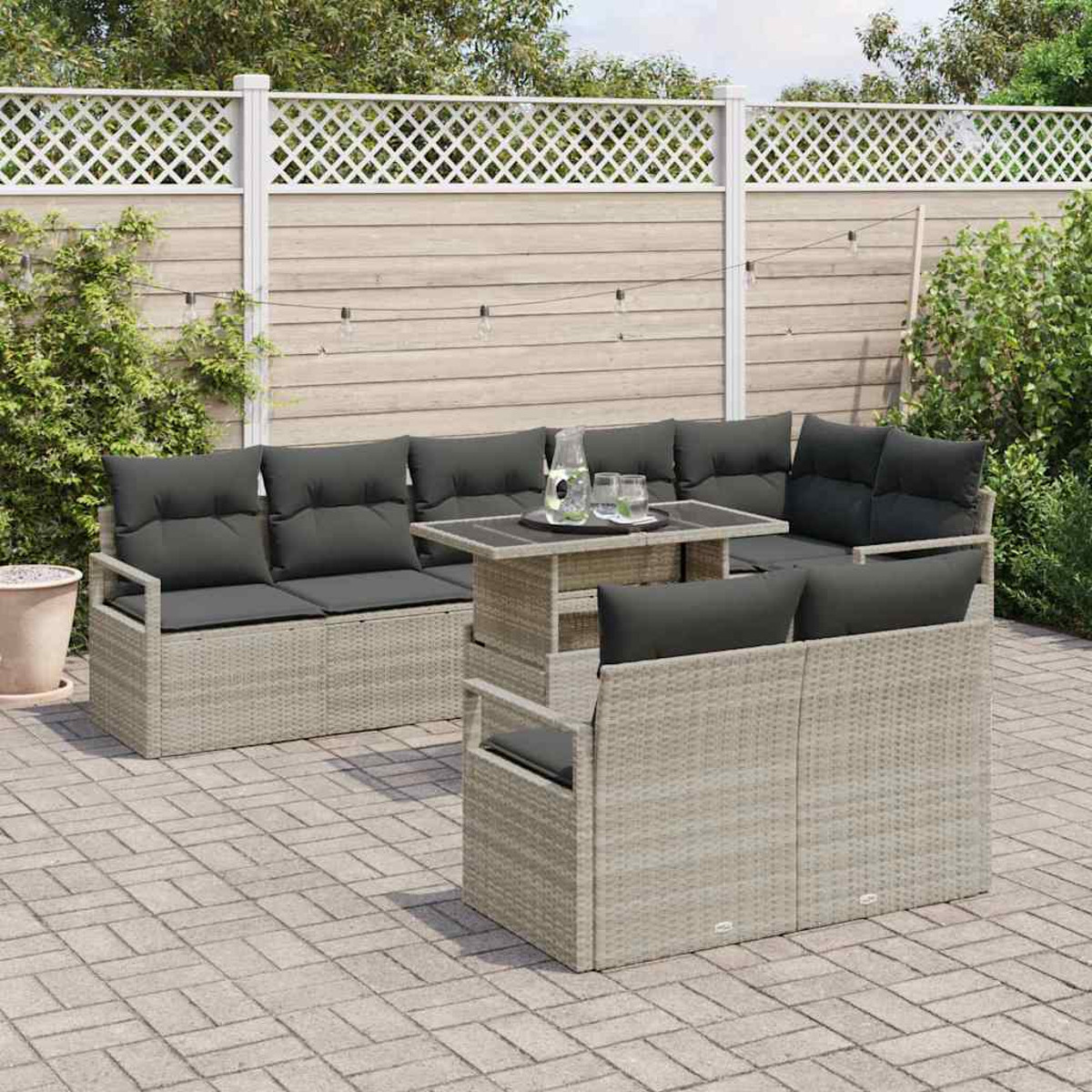 Gartenmöbel-Set aus Korbgeflecht mit grauen Kissen und einem kleinen Tisch. Das Set steht auf einer Steinterrasse, mit weißem Zaun und Grün im Hintergrund.