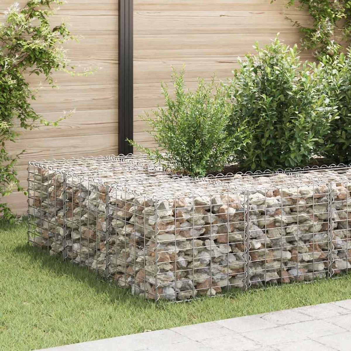 Ein mit Steinen gefüllter Gabion, der Pflanzen enthält. Es steht auf einem grünen Rasen. Holzzaun ist der Hintergrund.