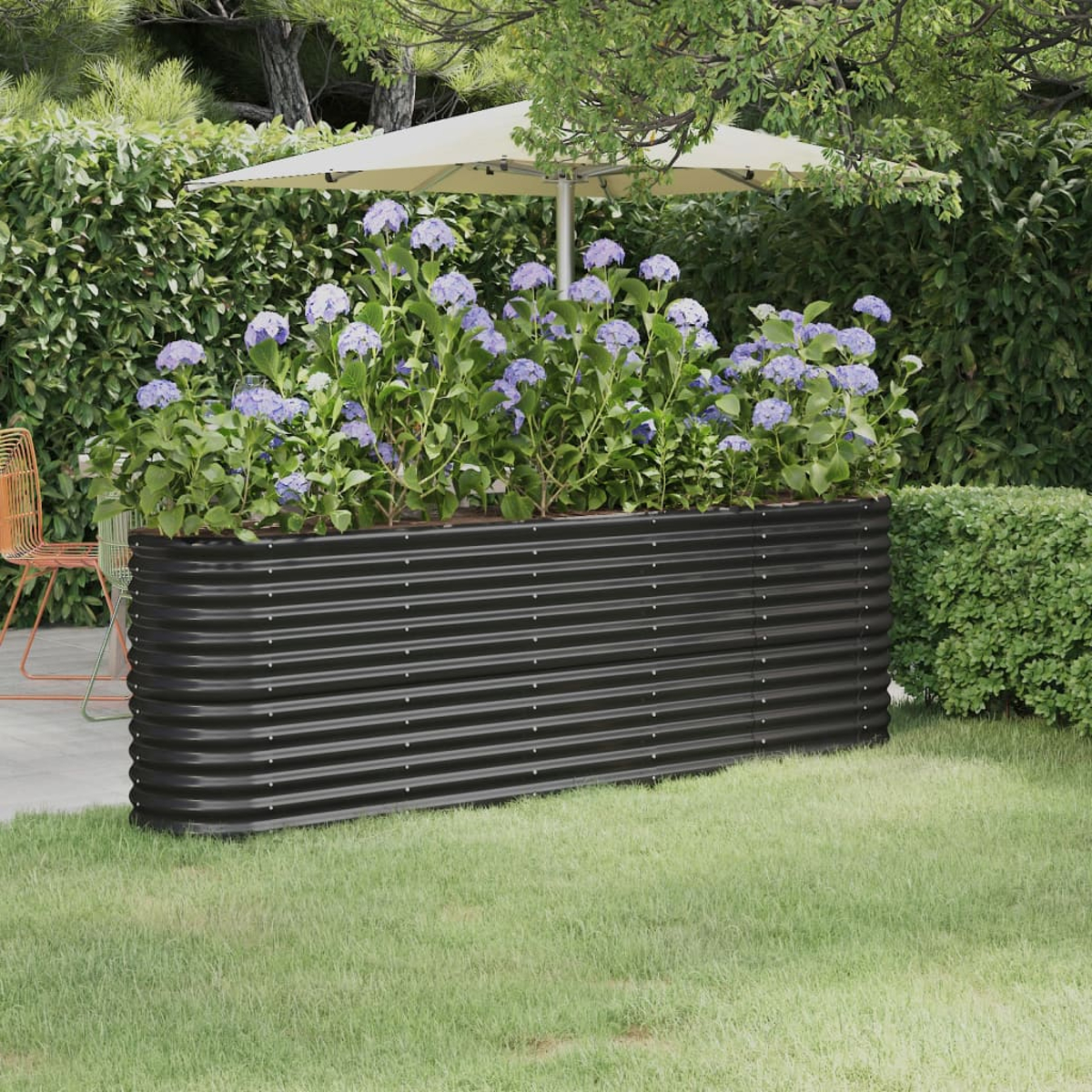 Ein schwarzer, gewellter Metall-Pflanzkasten mit lila Blumen steht auf Gras. Im Hintergrund befinden sich eine Terrasse, ein Schirm und Grün.