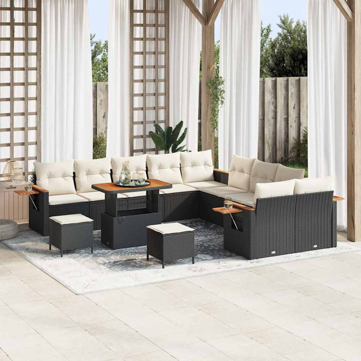 Set da patio esterno con mobili in vimini scuro, cuscini beige, un tavolo in legno e tende bianche. Piante verdi e luce solare.