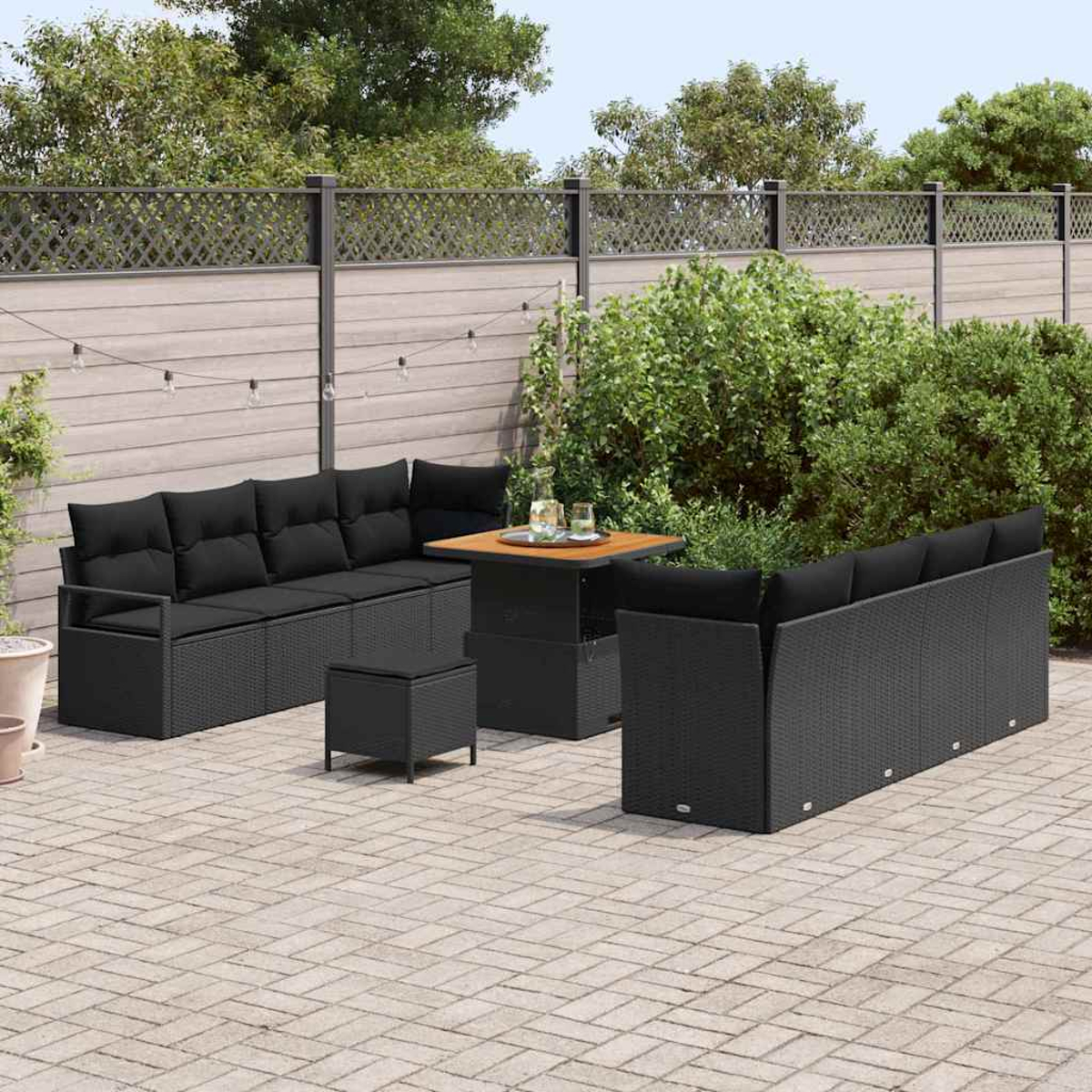 Terrassengarnitur im Freien mit schwarzen Korbs Sofas und einem Tisch. Vor einem Holzzaun mit dekorativem Oberteil befindet sich eine Steinterrasse.