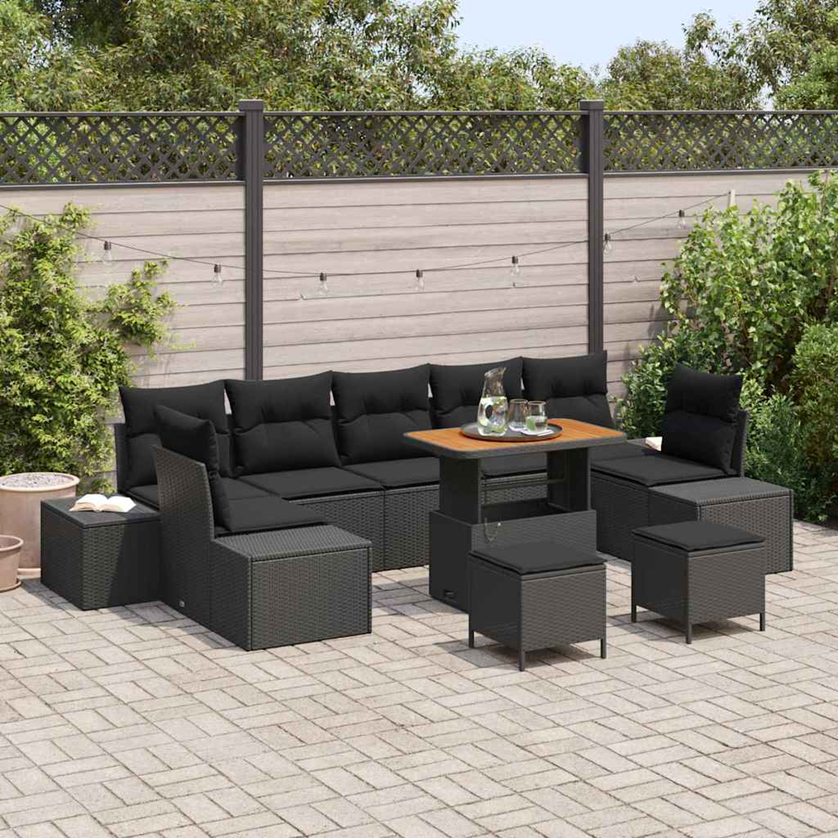 Terrassenset im Freien mit schwarzen Kissen, einem Holztisch und passenden Hockern. Die Szene spielt sich auf einer gepflasterten Terrasse ab.