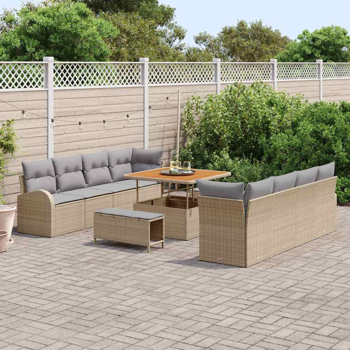 Set di mobili da esterno in vimini su un patio in mattoni. Include divani con cuscini grigi, un tavolo e una panca. Lo sfondo è una recinzione e alberi.