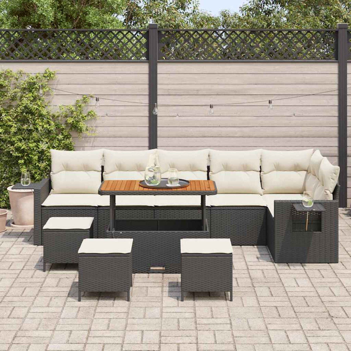 Outdoor-Terrassenset: dunkelgraues Korbmöbel-Sofa mit cremefarbenen Kissen, einem Holztisch und drei Hockern. Vor einem Holzzaun.