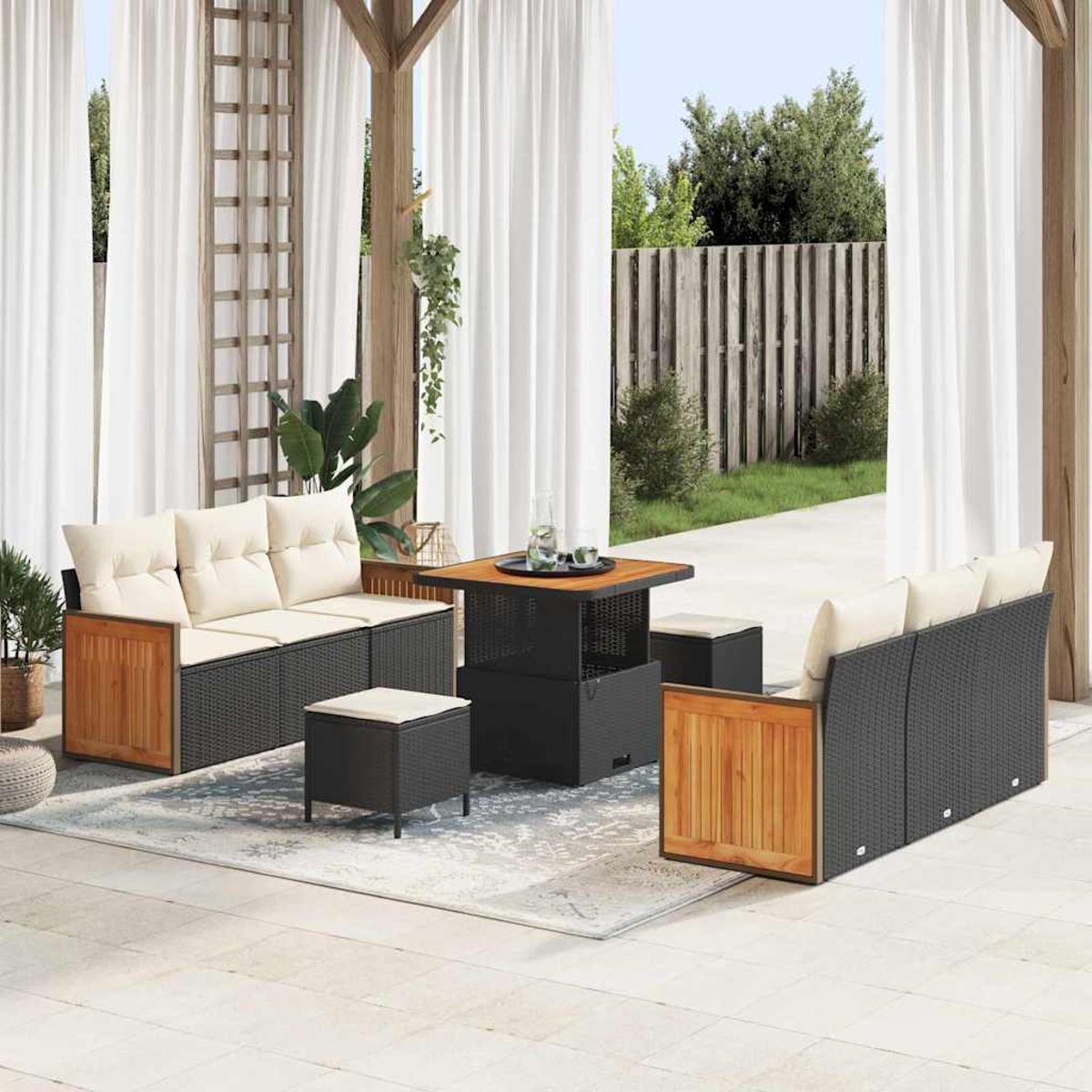 Set patio esterno con divani in rattan nero, dettagli in legno, cuscini color crema e un tavolo quadrato. Tende bianche e una staccionata in legno sullo sfondo.