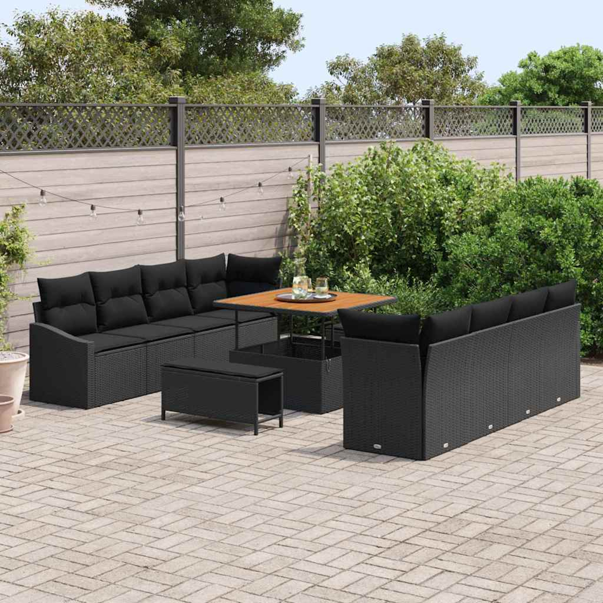 Terrassenmöbel-Set mit schwarzem Korbmöbel, einem Holztisch und einer Bank. Grüne Büsche und ein Zaun im Hintergrund.