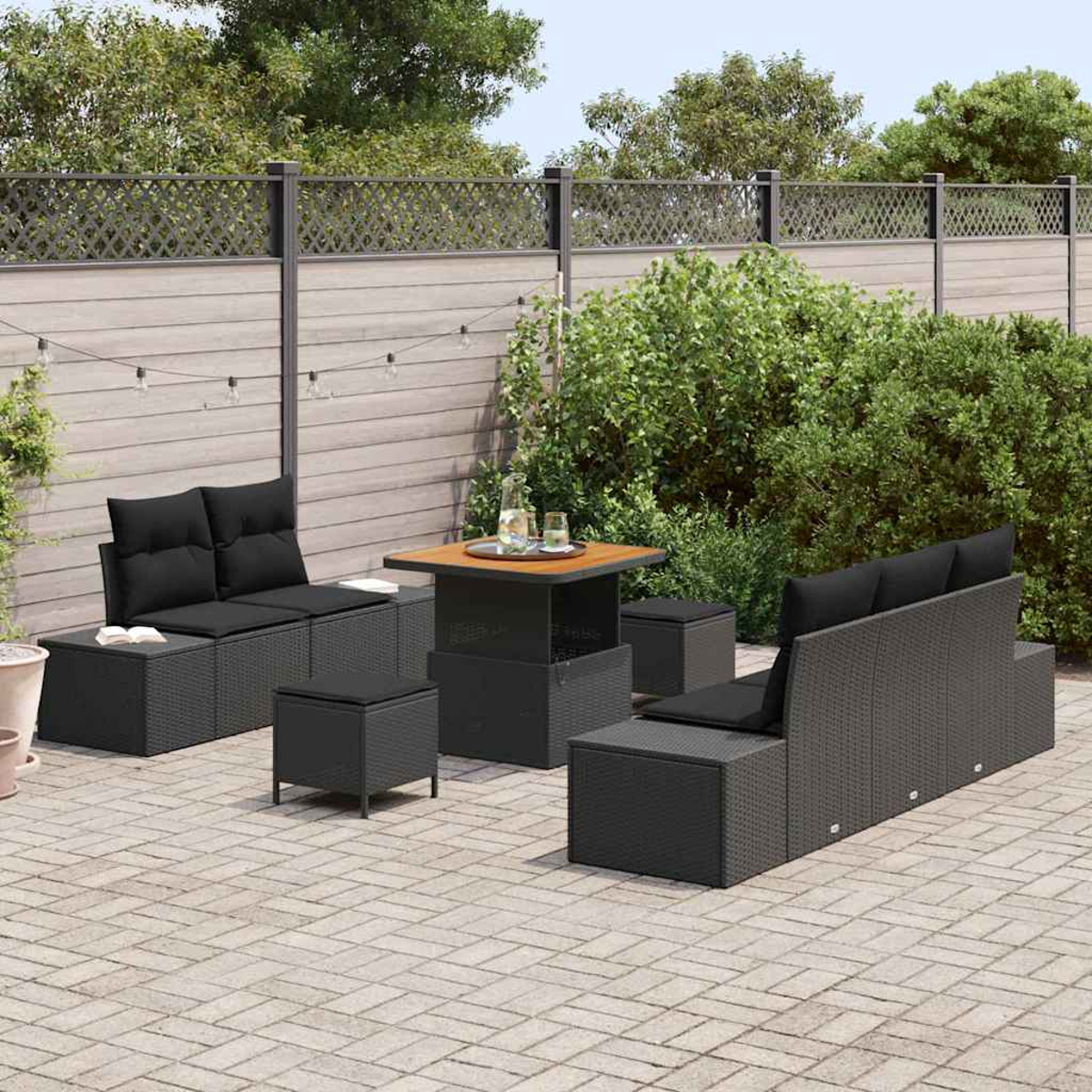 Garten-Terrassenset. Schwarze Korbmöbel mit Kissen, Tisch und Hockern auf einer Steinterrasse, mit Holzzaun und Grün.