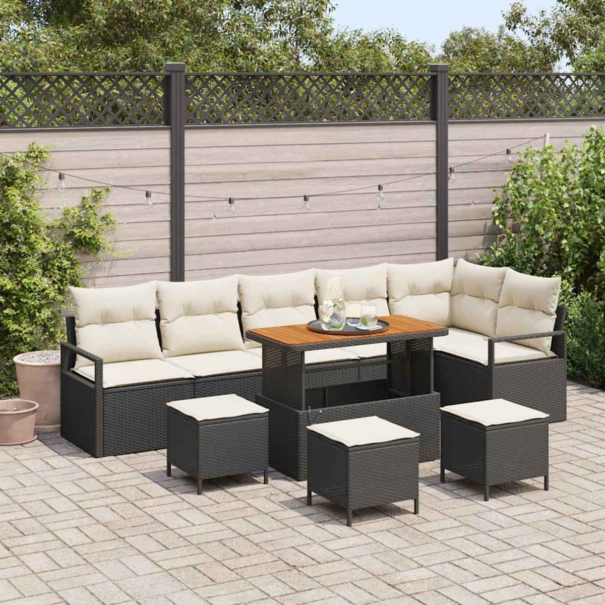 Garten-Terrassenset mit dunkelgrauem Korbsessel, Tisch und Hockern. Beige Kissen. Tischplatte aus Holz. Hintergrund mit Zaun und Pflanzen.