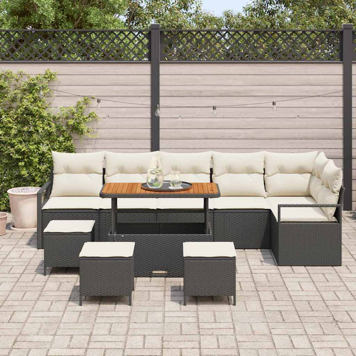 Outdoor-Ecksofa mit cremefarbenen Kissen, kleinem Holztisch und Ottomanen auf einer Terrasse, vor einem Holzzaun mit Pflanzen.